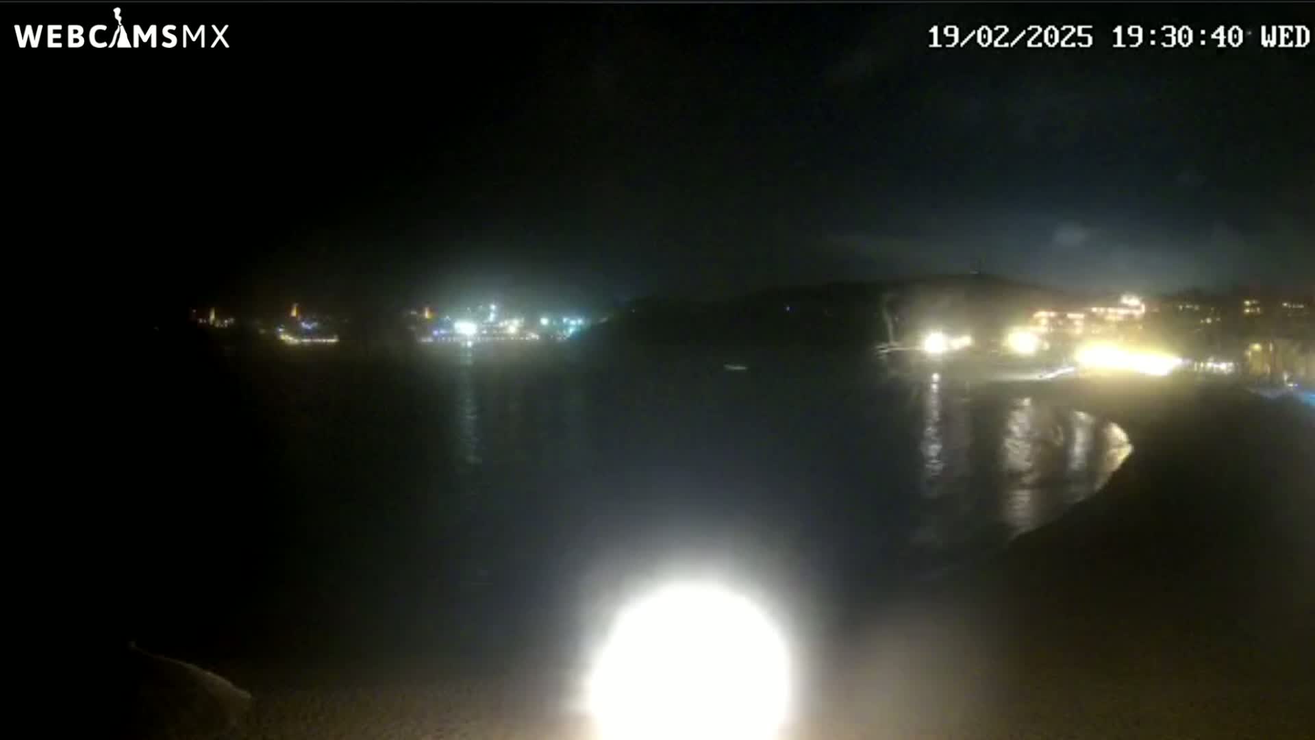 A nighttime, seaside view shows a dark body of water reflecting the lights from buildings on the opposite shore.
