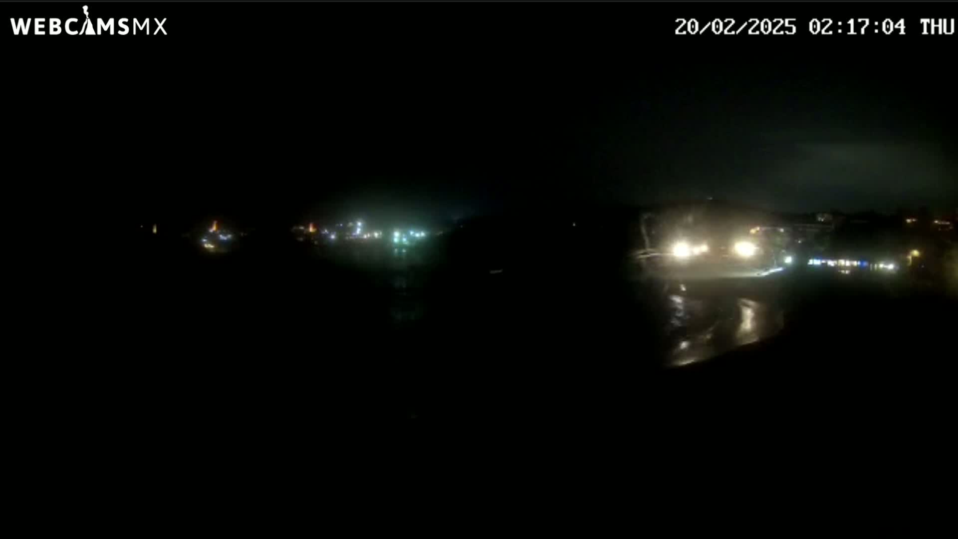 A nighttime, rain-slicked road is visible with a few vehicles driving past, showing some distant lights in the background.