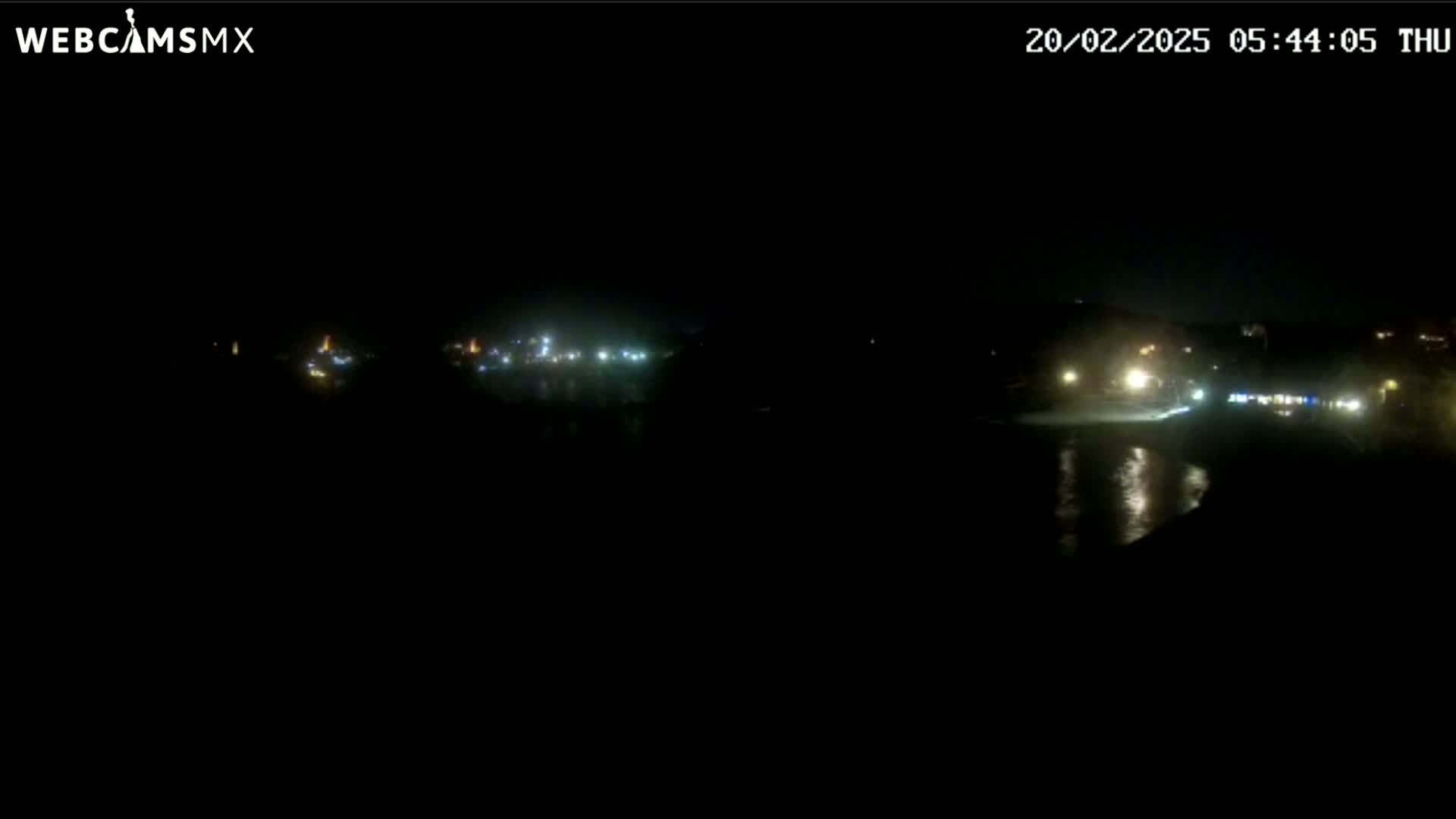 A dark nighttime scene shows a body of water reflecting distant lights from a shoreline at night.