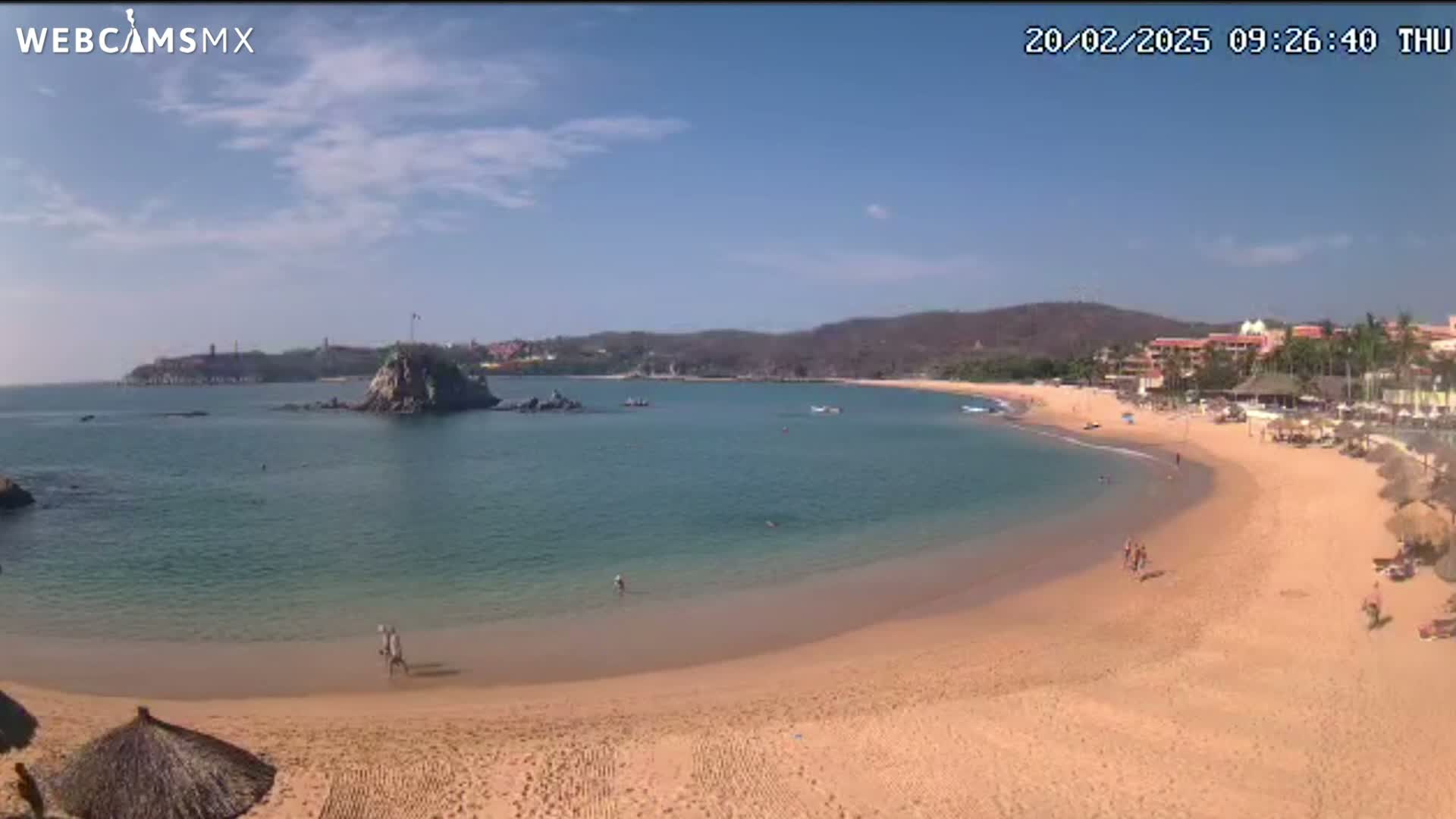 A sandy beach curves around a calm bay with a rocky island and distant buildings under a mostly sunny sky.