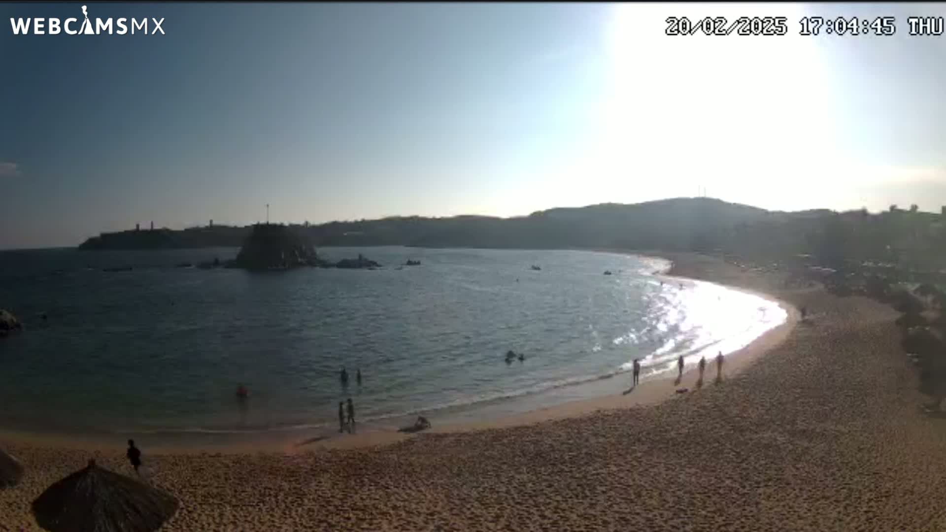 A sandy beach curves around a calm bay on a sunny day, with a few people swimming and walking along the shore.
