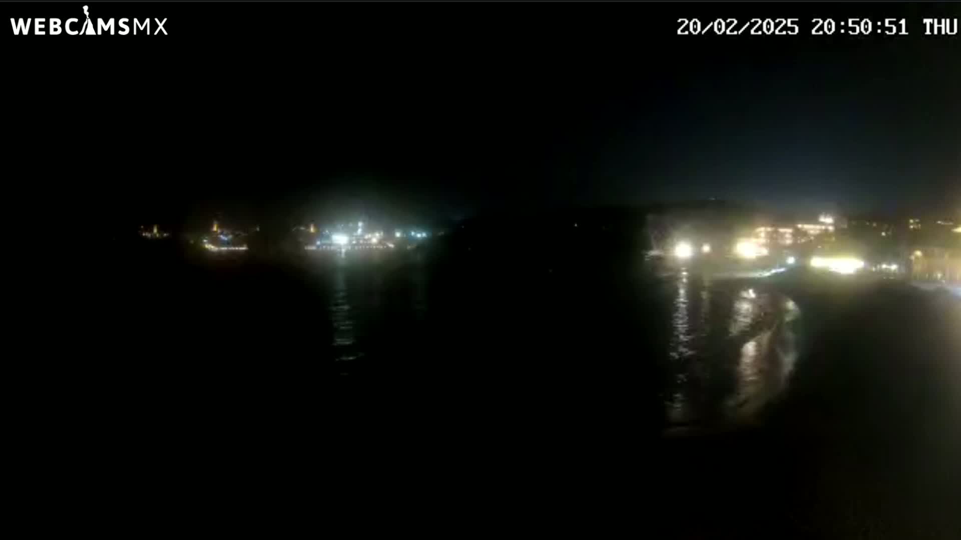 A nighttime view of a dark body of water reflecting the lights of buildings on the shore under a clear sky.