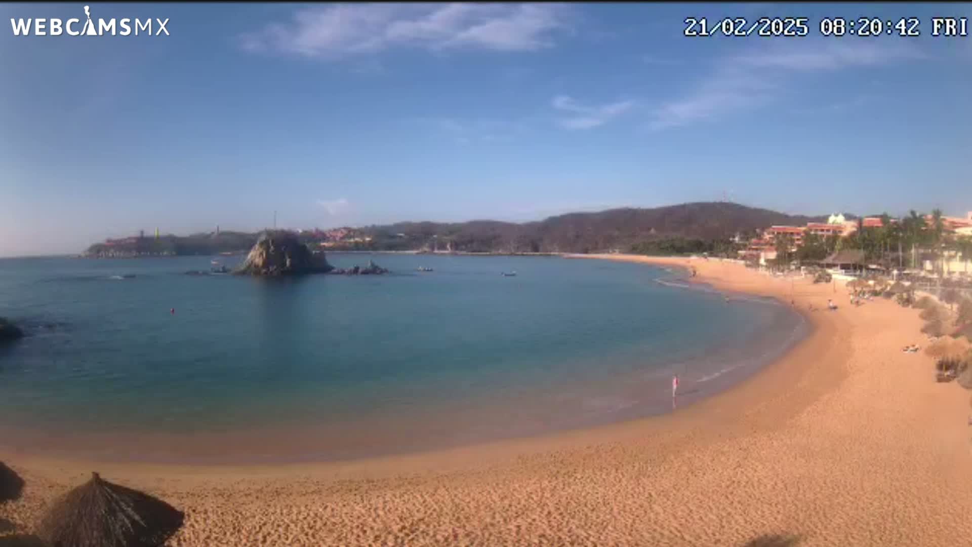 A secluded cove with a sandy beach and calm, clear turquoise water under a mostly sunny sky is seen from an elevated vantage point.