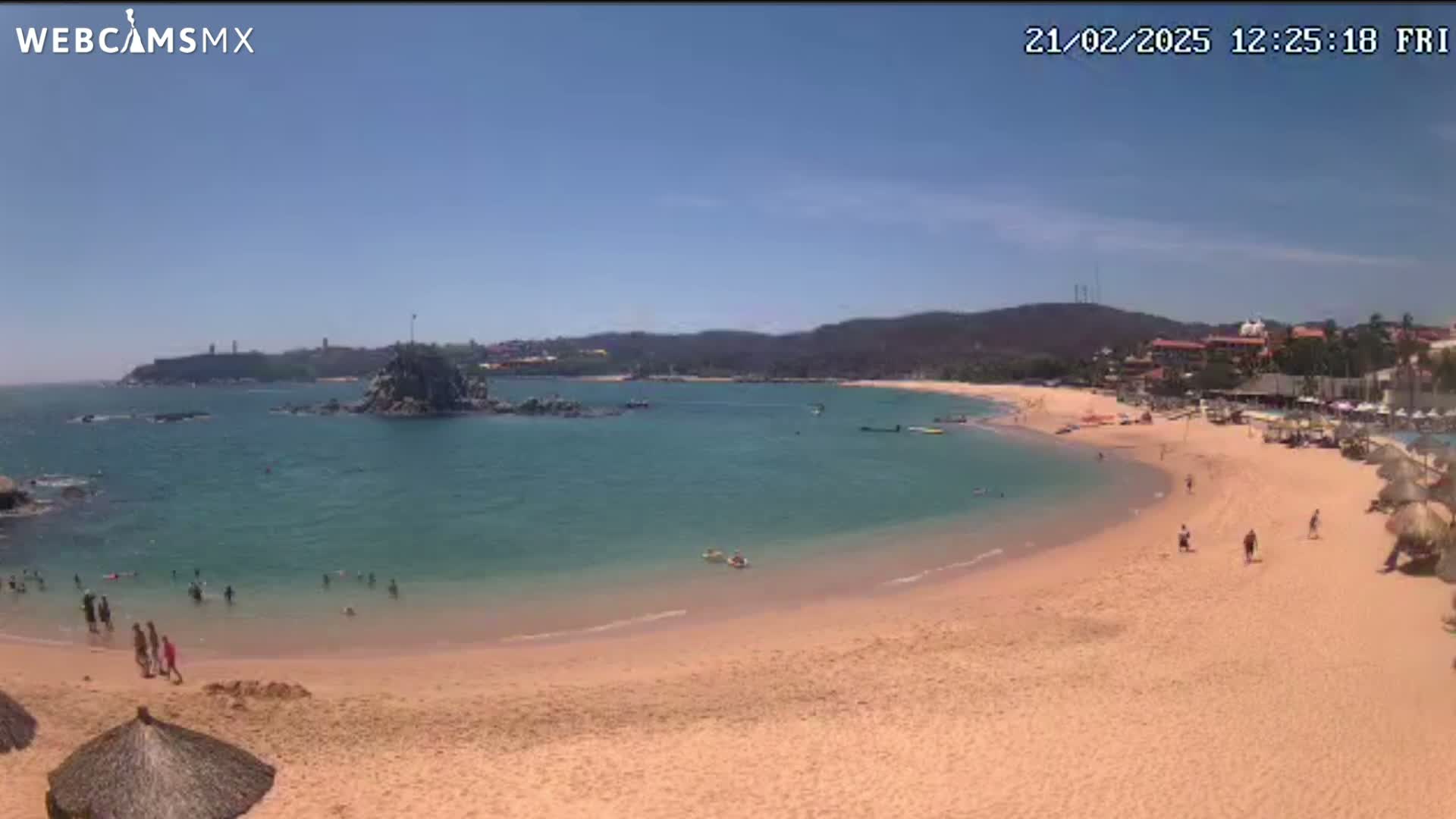 A sandy beach curves around a calm, clear bay under a bright, sunny sky, with people swimming and relaxing on the shore and in the water.