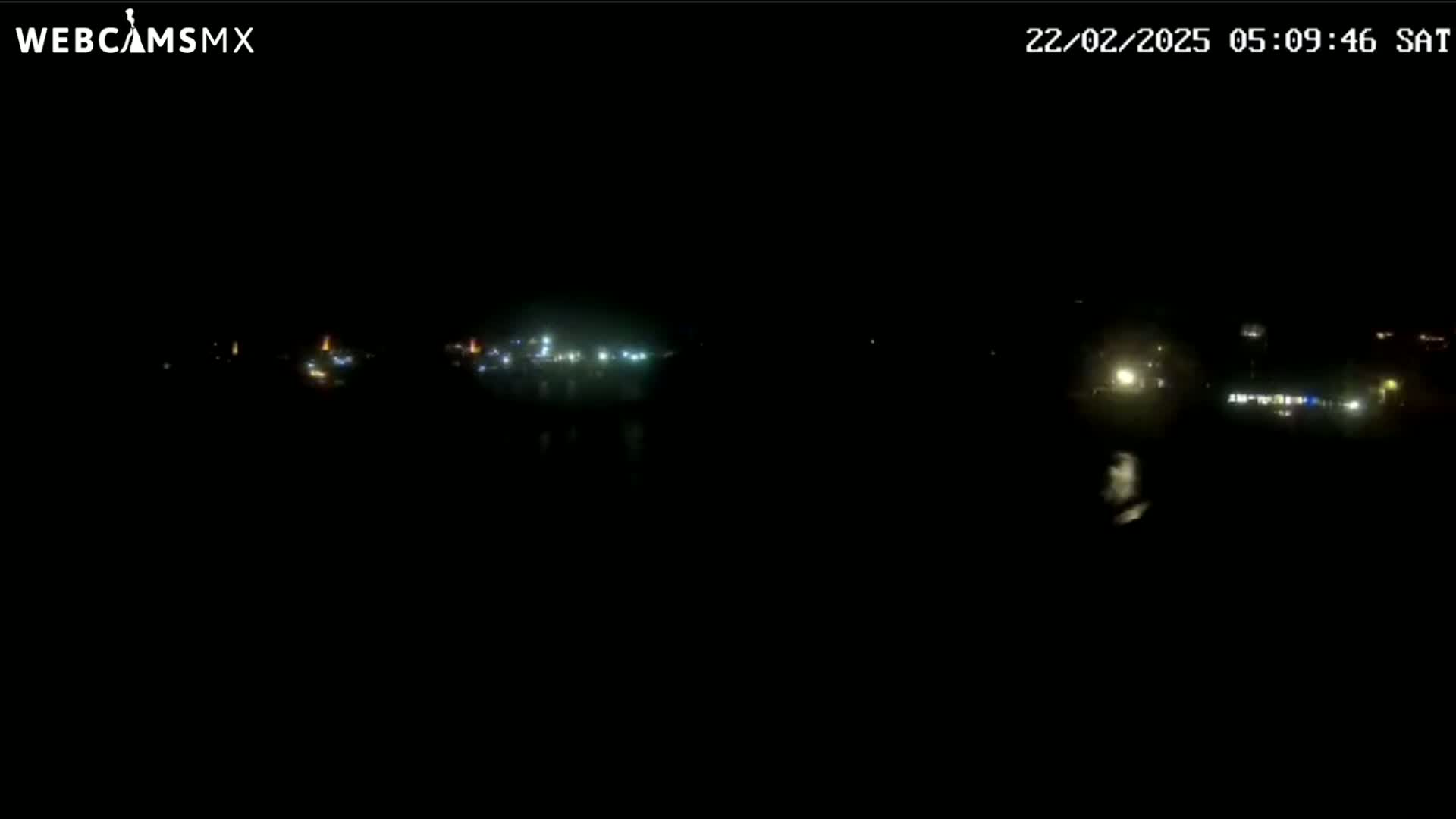 A nighttime, long shot shows a dimly lit waterfront area with scattered lights visible in the distance.