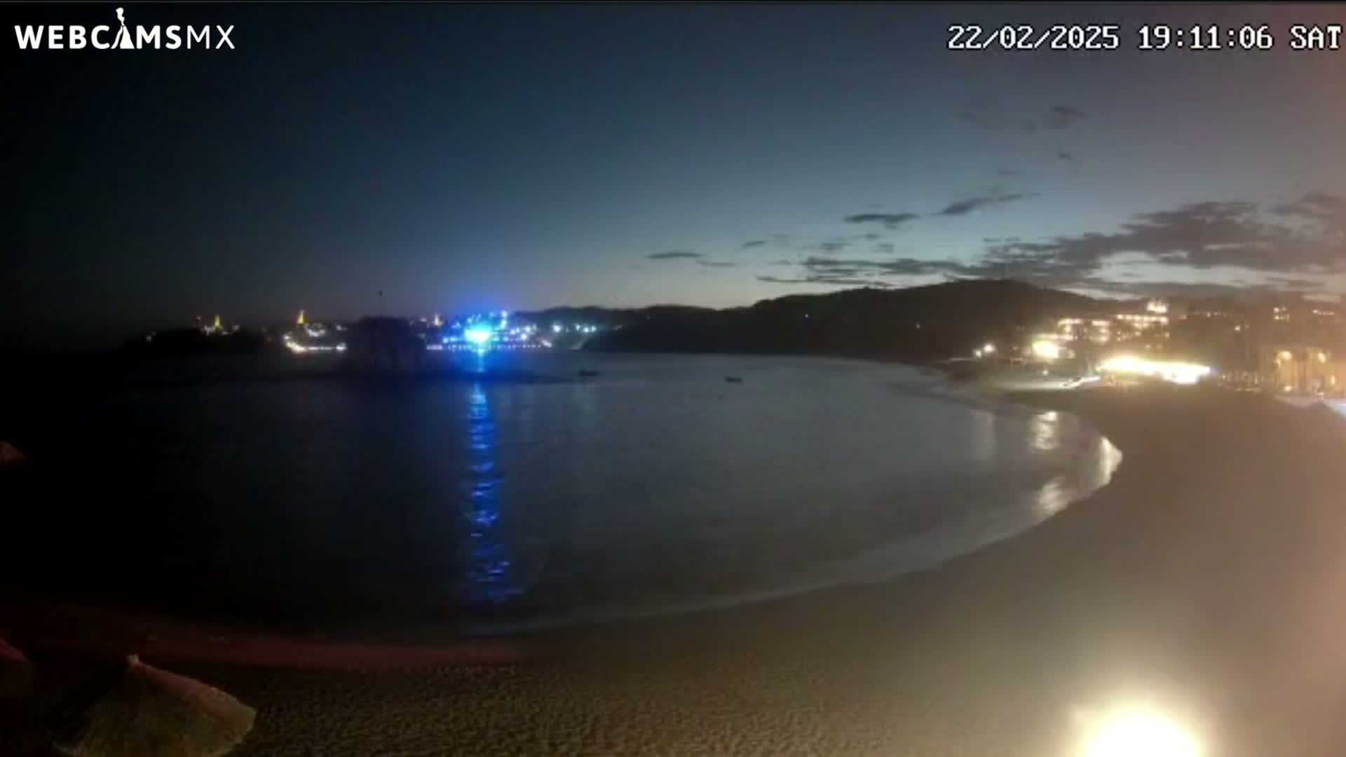 A nighttime view of a dimly lit beach with calm water, and buildings with lights across the bay under a mostly clear, dark sky.