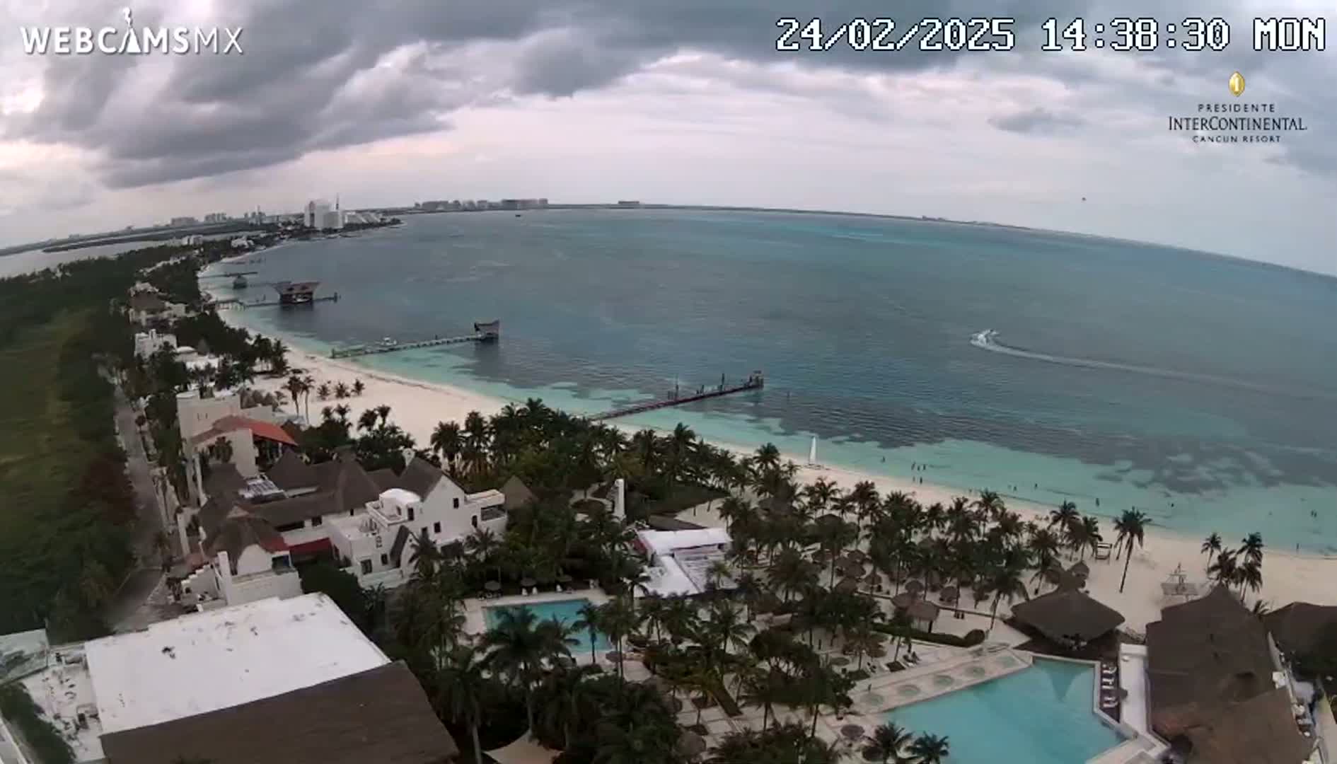An aerial view shows a tropical beach with resorts, palm trees, and calm turquoise water under a cloudy sky.