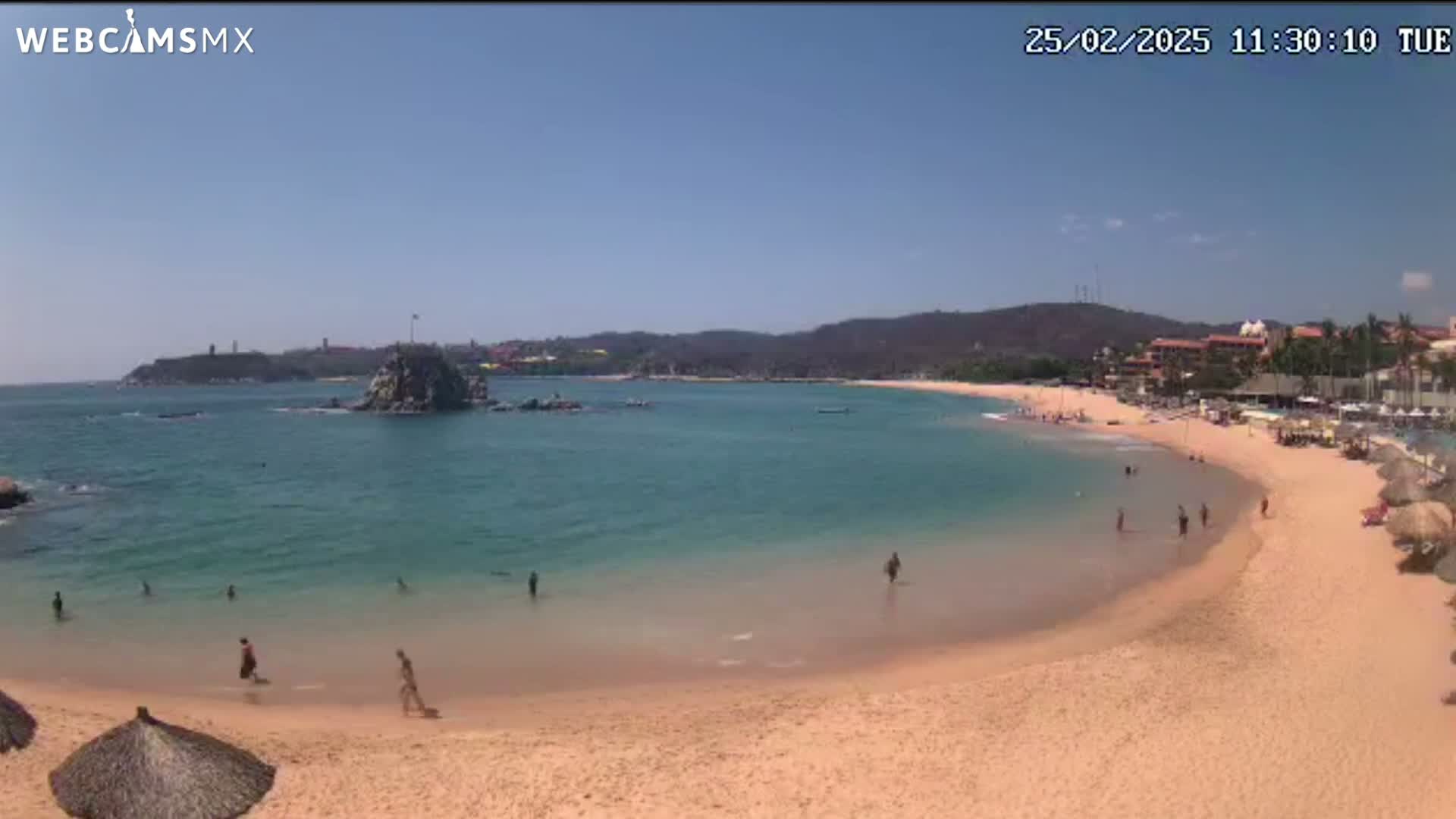 A sunny beach with calm, clear turquoise water curves around a rocky outcropping, with a few people swimming and relaxing on the sand.