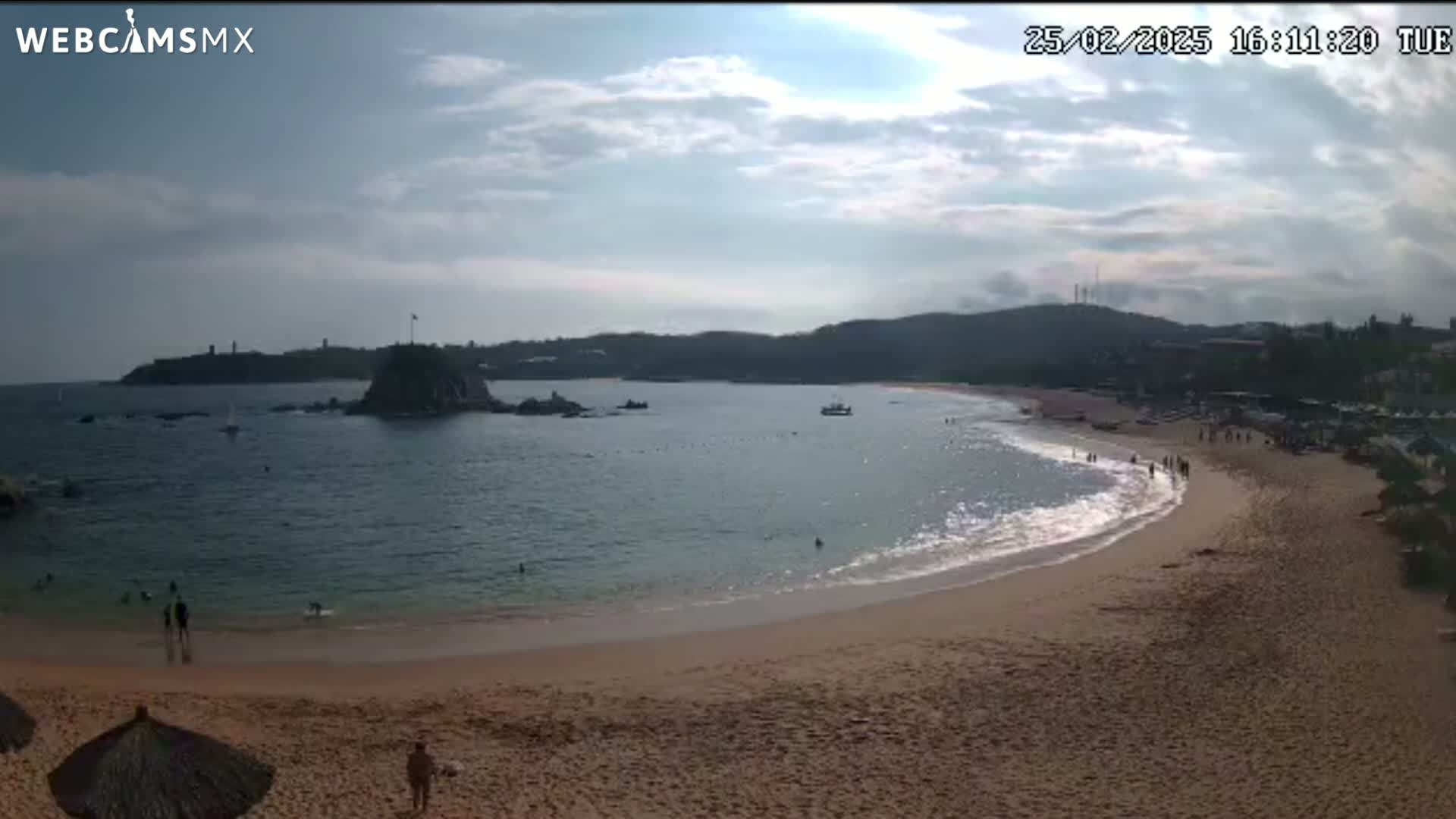 A partly cloudy day shows a sandy beach curving into a calm bay with a rocky island and distant hills.