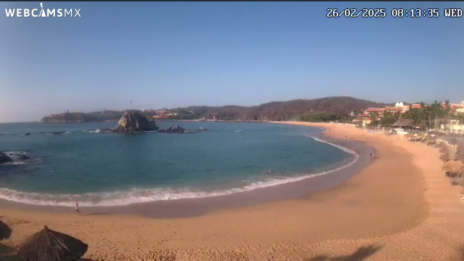 A sandy beach curves into a calm bay of blue water under a clear sky, with a rocky outcropping and distant buildings visible.