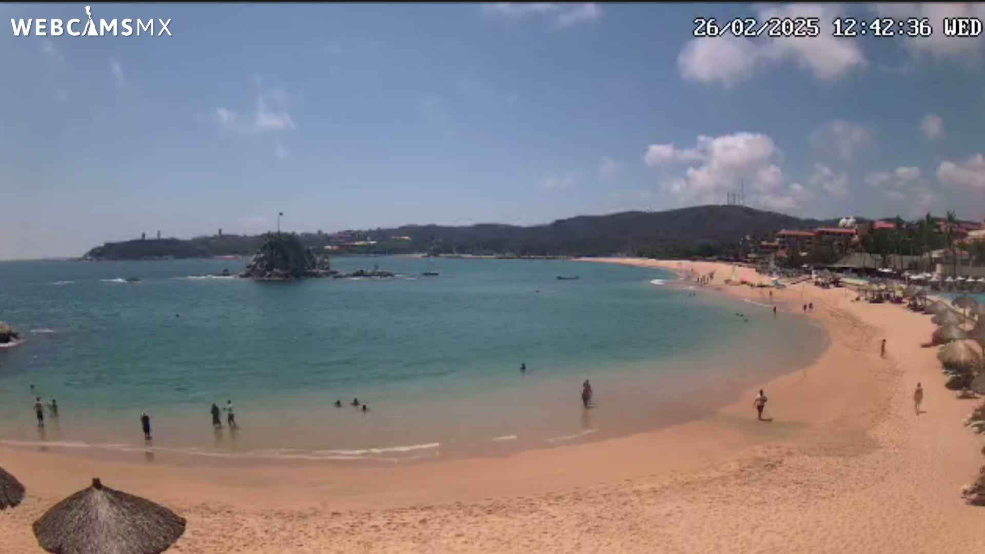 A partly sunny day at a beach with calm, clear turquoise water, a sandy shore, and a few people swimming and relaxing.