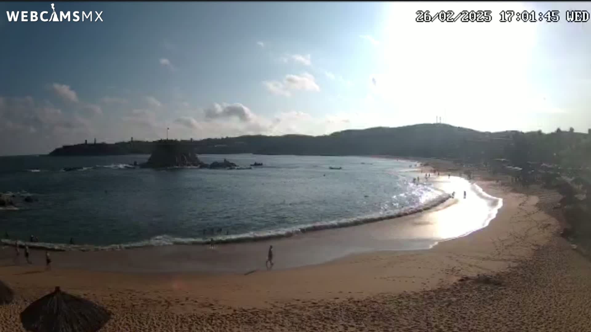 A sandy beach curves around a calm bay, with a rocky island and hills visible in the background under a partly sunny sky.