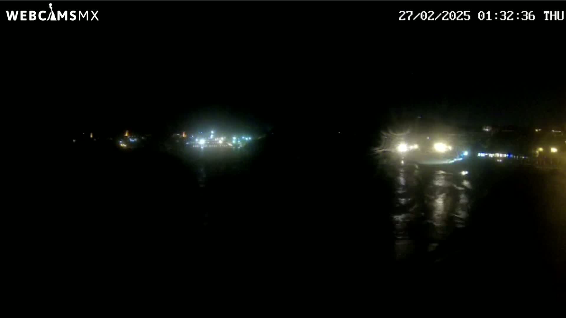 A nighttime, wet road scene shows the headlights of approaching vehicles reflecting on a dark, wet surface, with distant lights illuminating a town in the background.