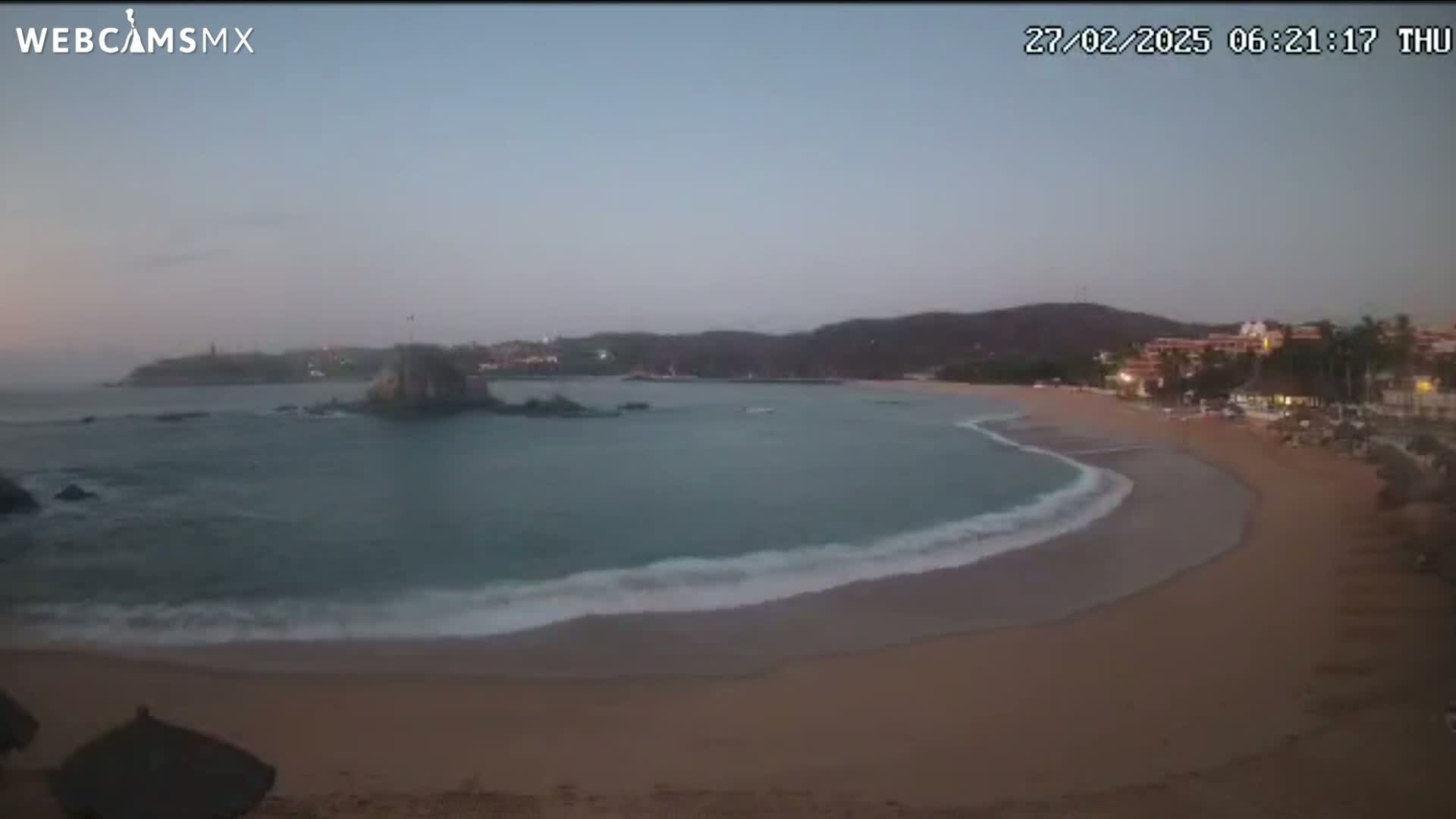 A sandy beach curves into a calm bay under a dusky sky, with small waves lapping the shore and a rocky island visible in the distance.