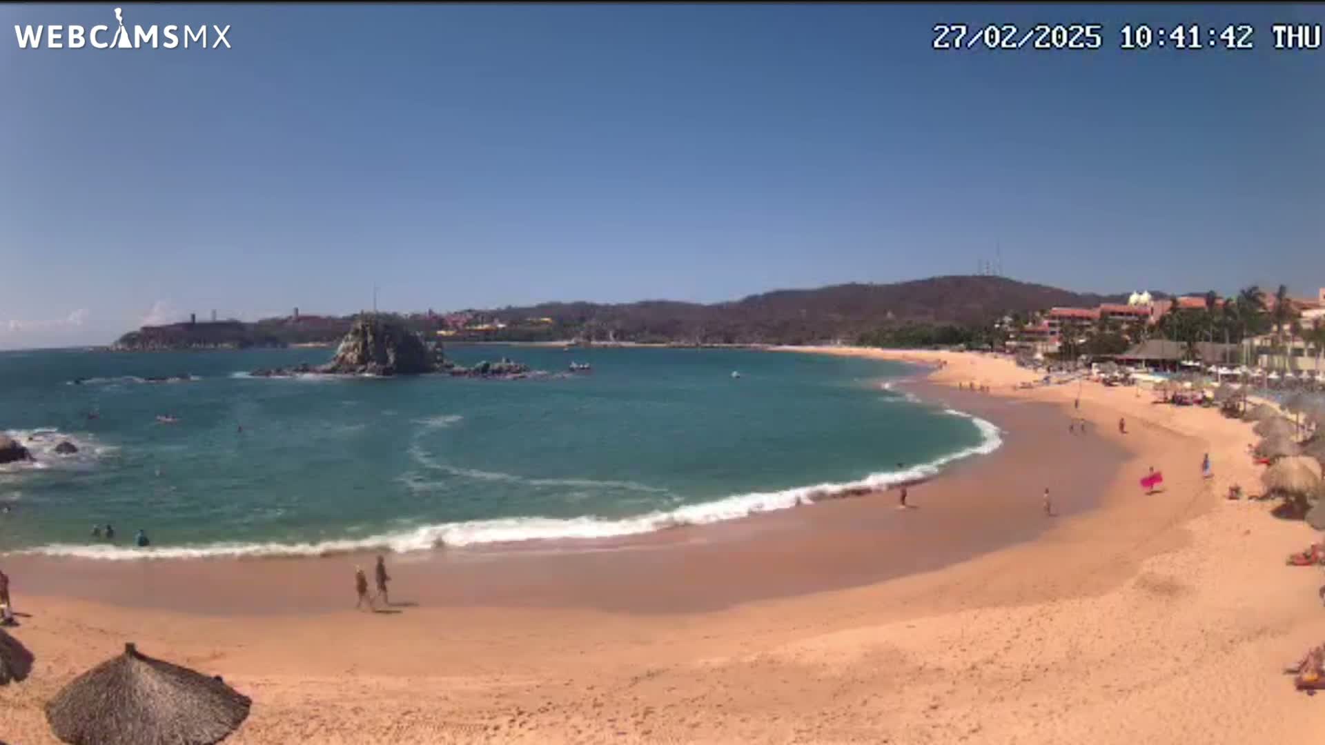 A sandy beach curves around a calm, turquoise bay under a clear blue sky, with a rocky islet and distant hills visible.