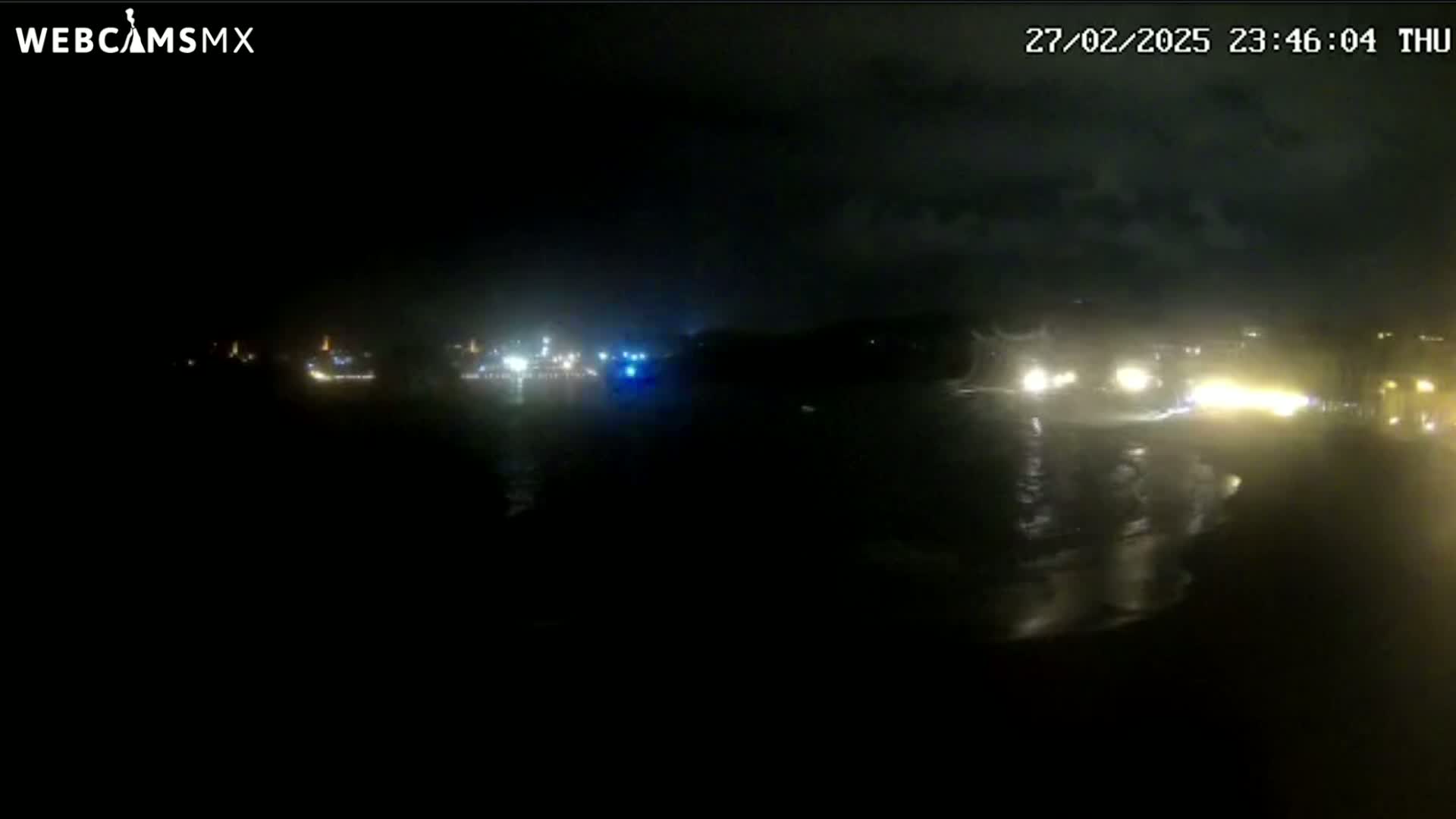 A nighttime, dark, and overcast scene shows faint lights from vehicles and buildings across a body of water.
