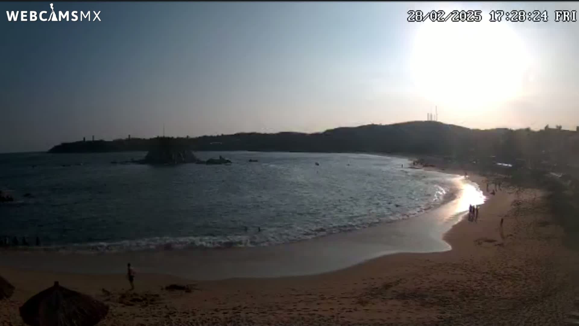 A sandy beach curves around a calm ocean cove at sunset, with a few people visible on the shore and in the water.