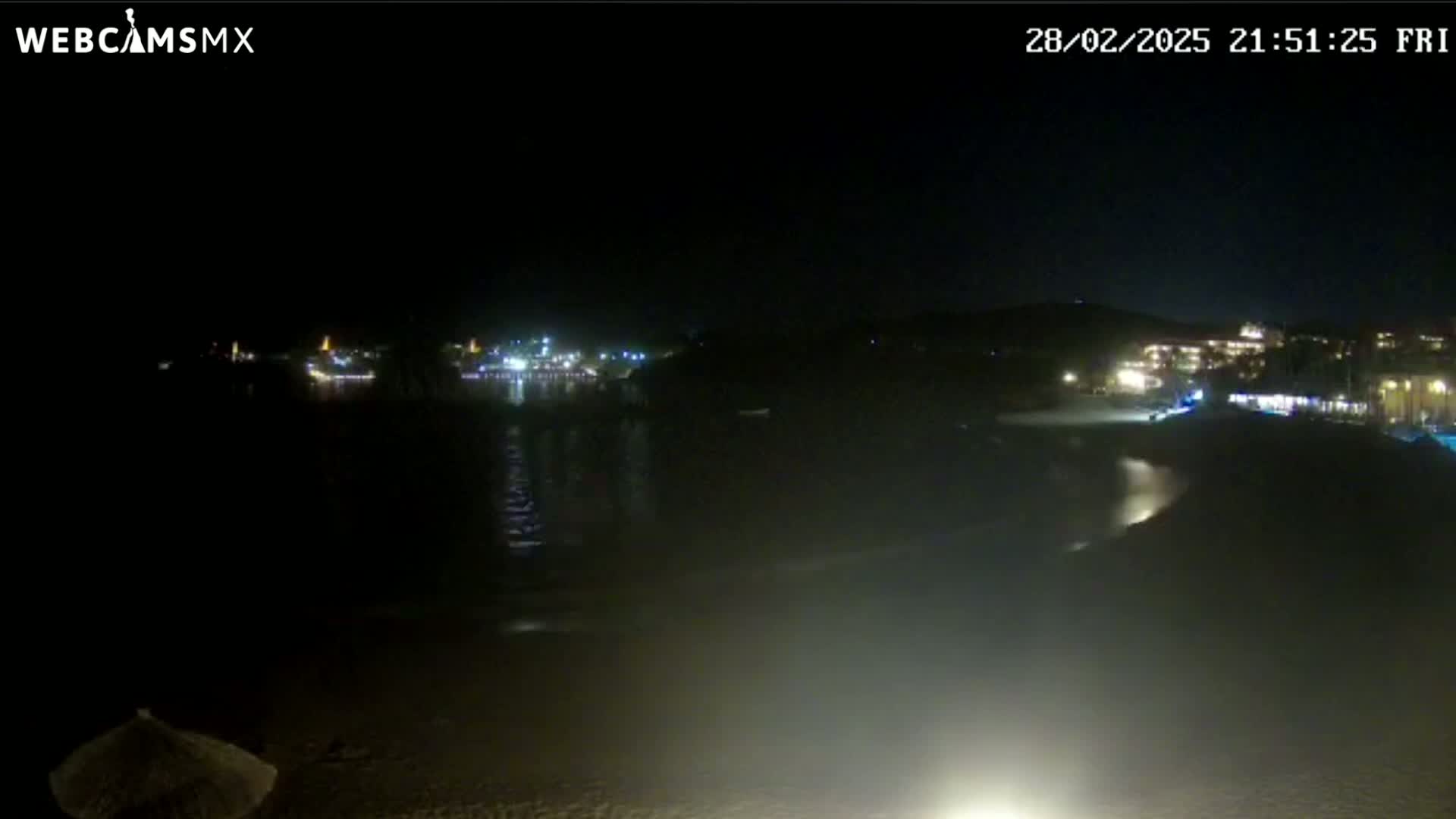 A dark, nighttime beach scene shows a calm body of water reflecting the lights of buildings across the water under a clear sky.