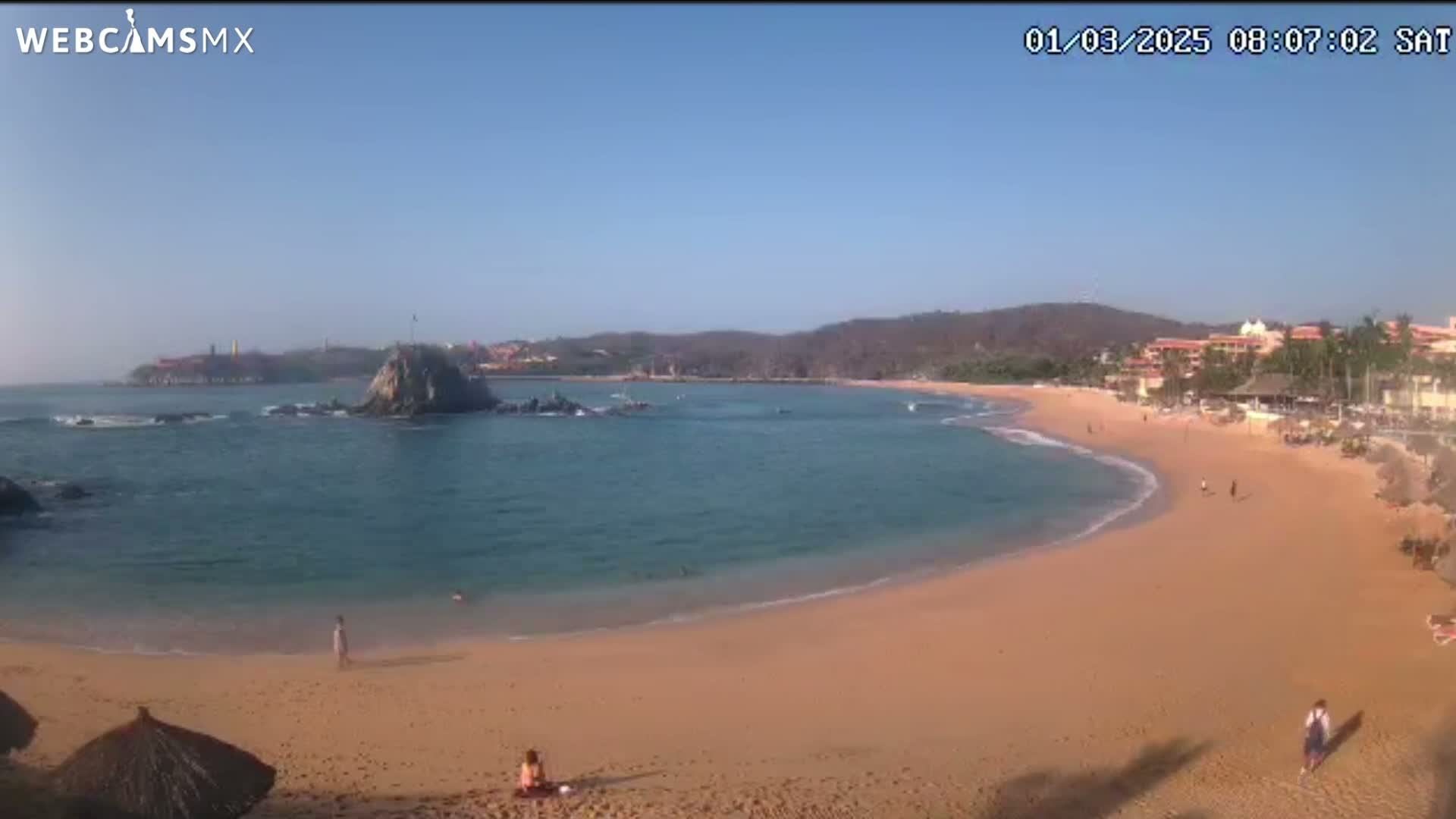 A sandy beach curves around a calm, blue bay under a clear blue sky, with a rocky outcrop and distant buildings visible.