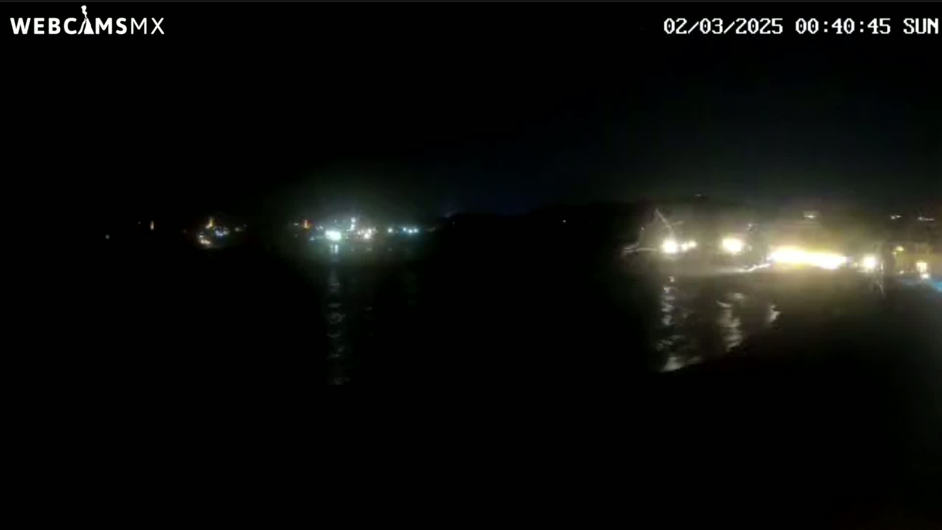 At night, vehicle headlights illuminate a dark body of water reflecting the lights of a distant shoreline.