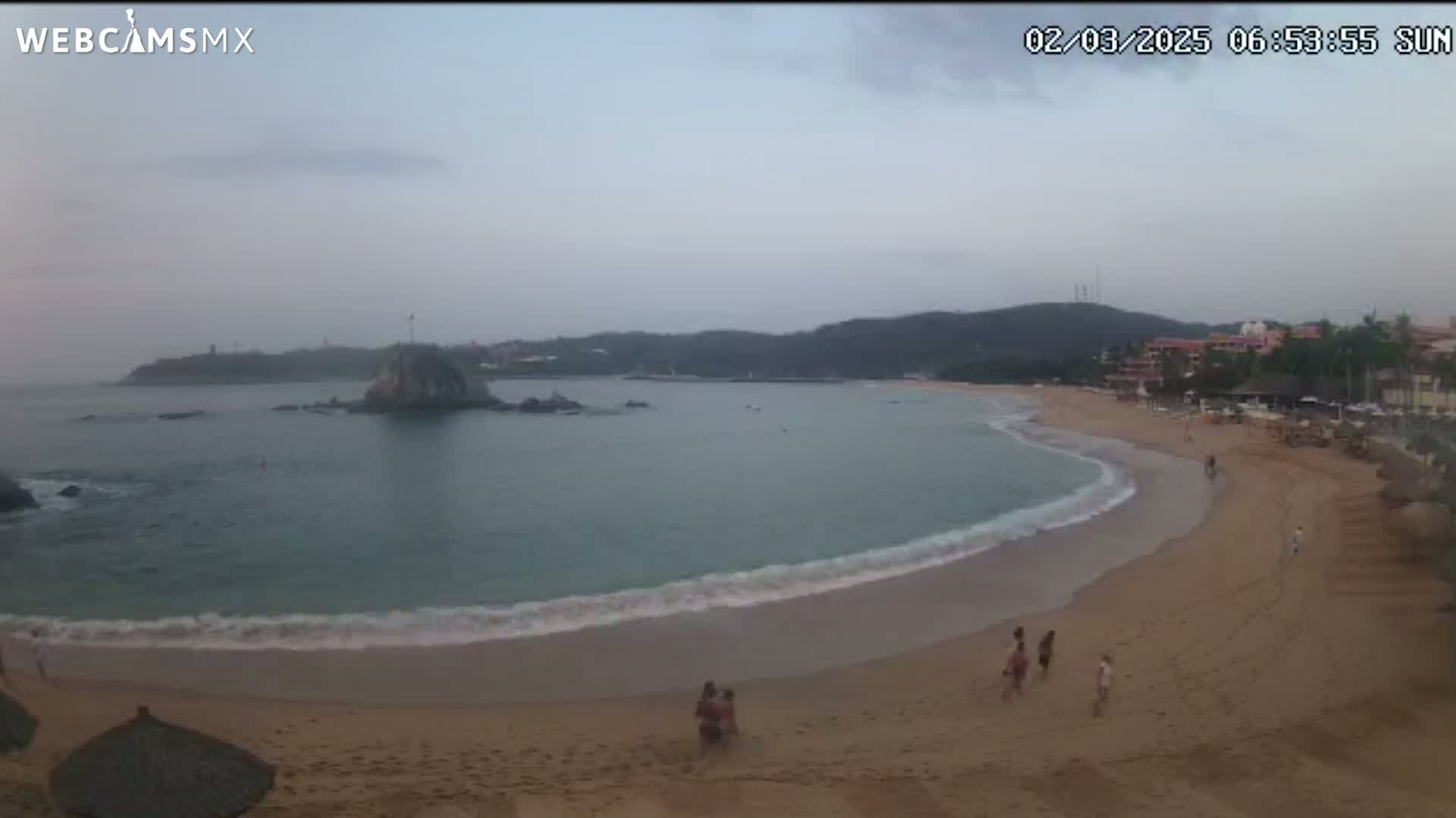 A sandy beach curves around a calm bay under an overcast sky, with a few people walking along the shore and a small island visible in the water.