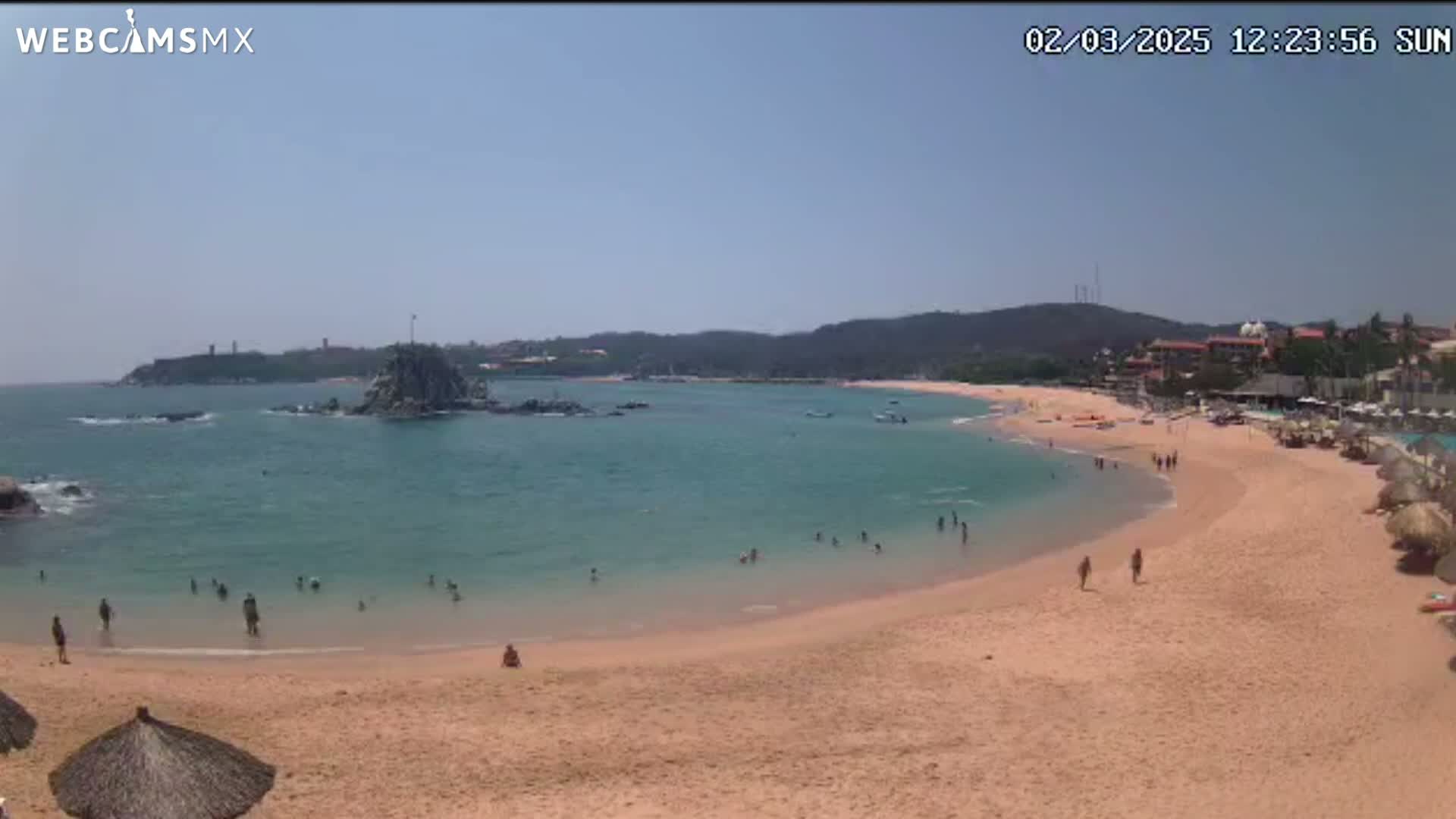 A sandy beach curves around a calm, turquoise bay under a clear, sunny sky, with people swimming and sunbathing.
