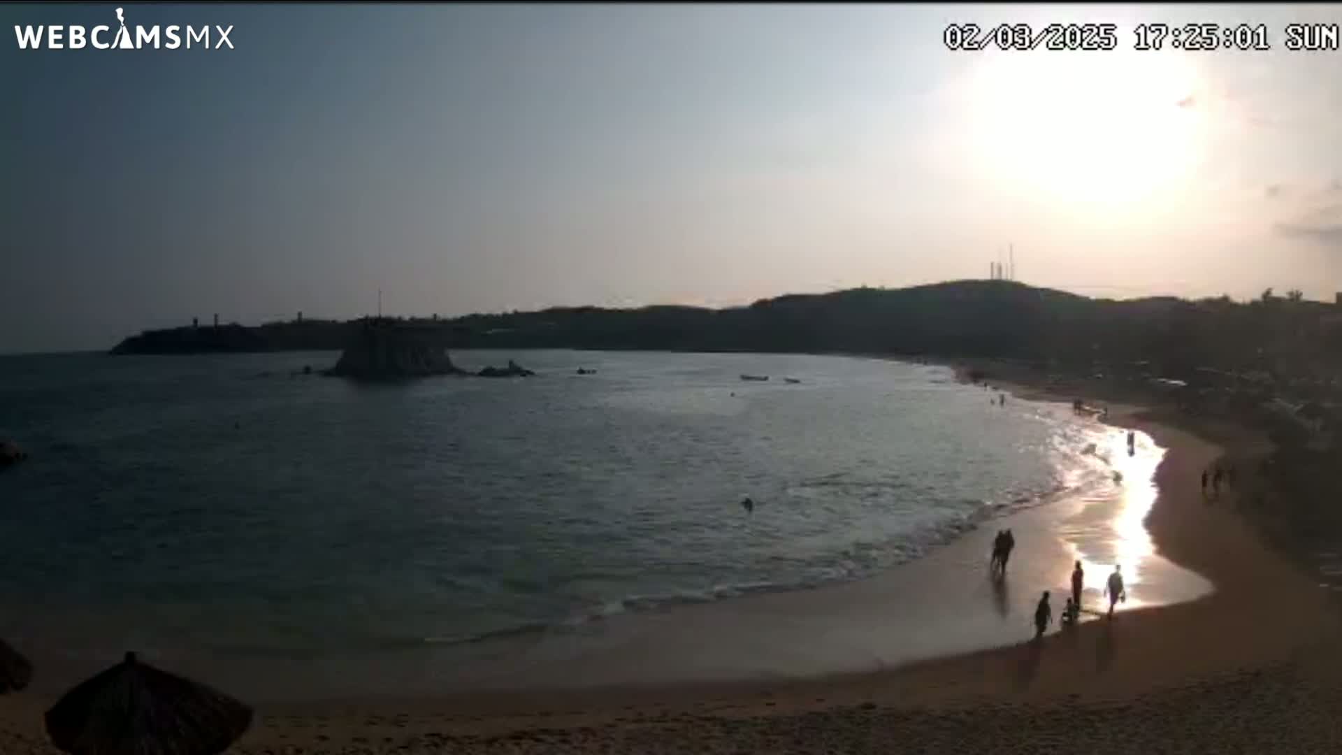 A beach scene at sunset shows calm ocean waves lapping a sandy shore where several people are walking, with a rocky island visible in the bay.