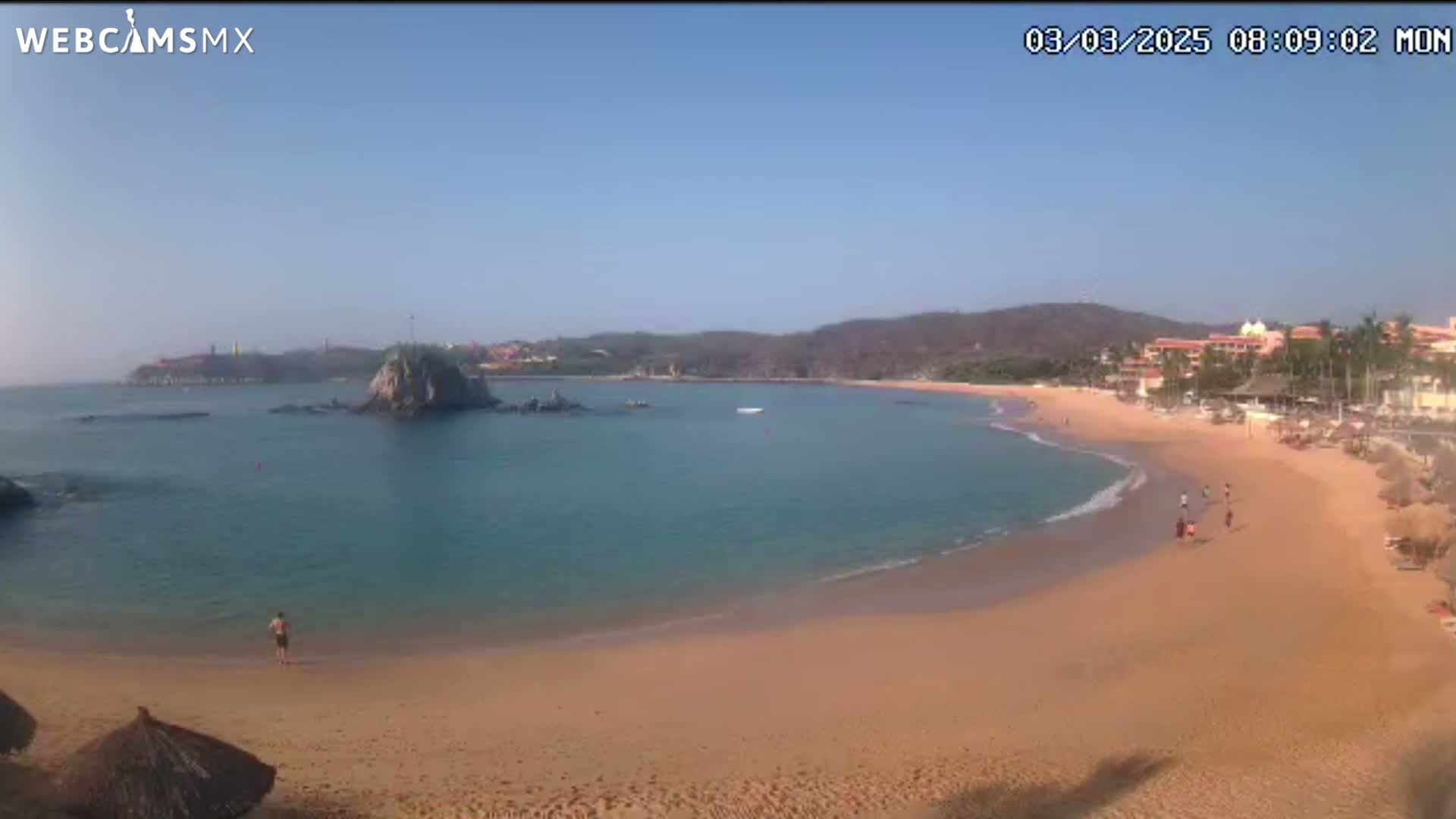 A secluded cove with a sandy beach, calm turquoise water, and a small rocky island is viewed on a clear, sunny day.