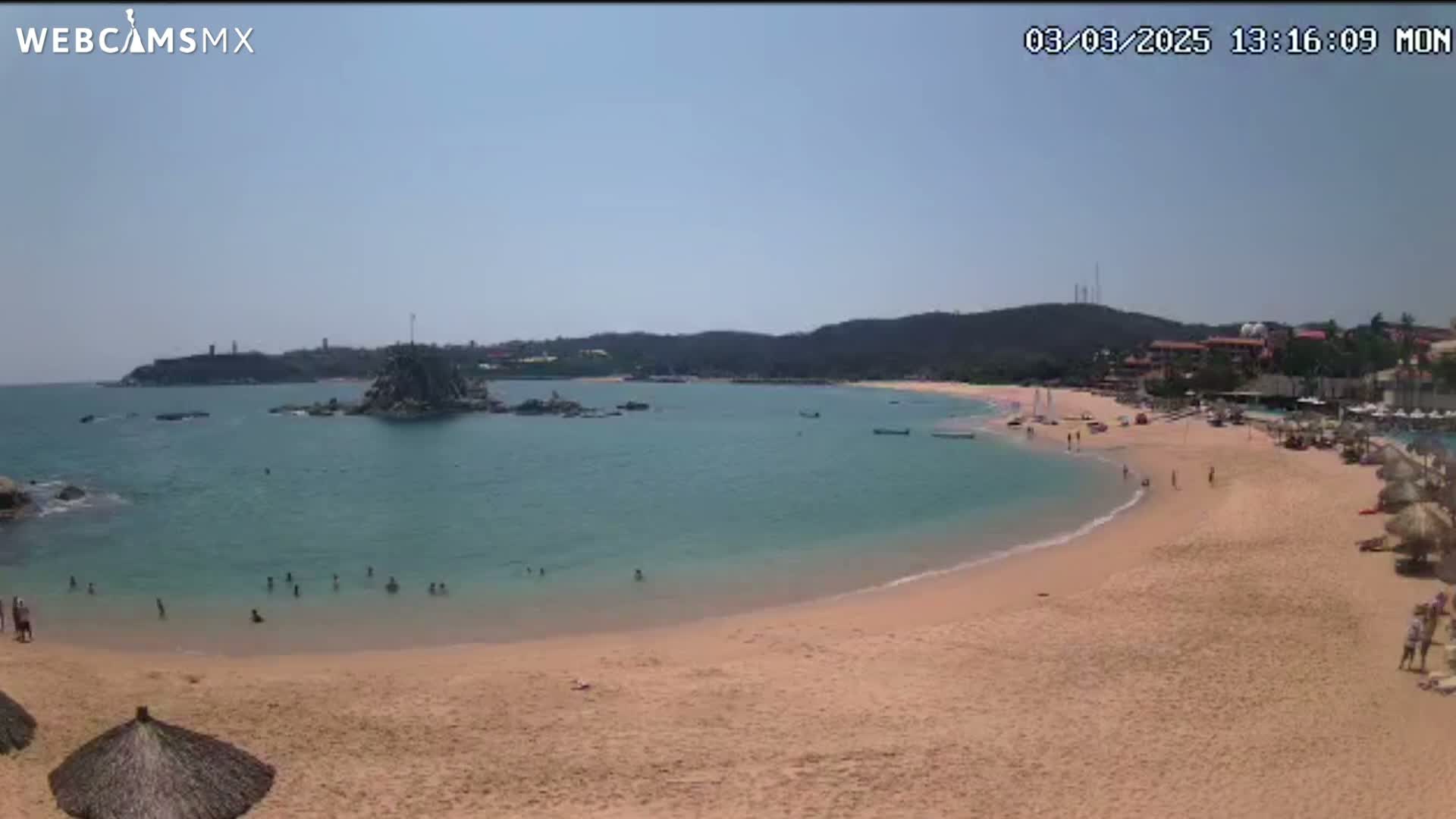 A sandy beach curves into a calm, teal ocean under a clear, sunny sky; several people are swimming and relaxing on the beach.