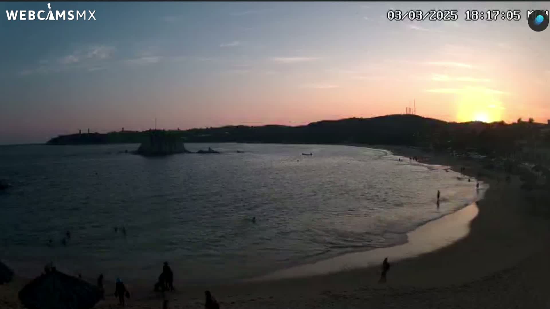 A beach scene at sunset shows calm water lapping a sandy shore, with a few people visible on the beach and in the water.