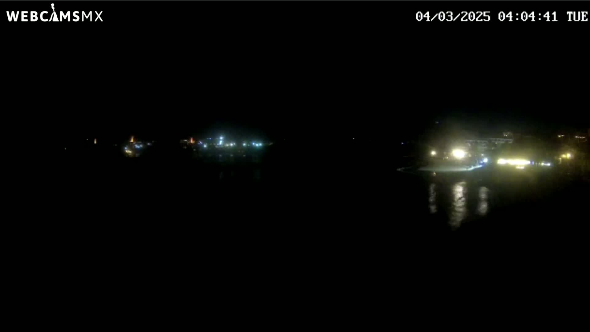 A nighttime, dark view of a body of water reflecting distant lights from buildings and structures on the shore.