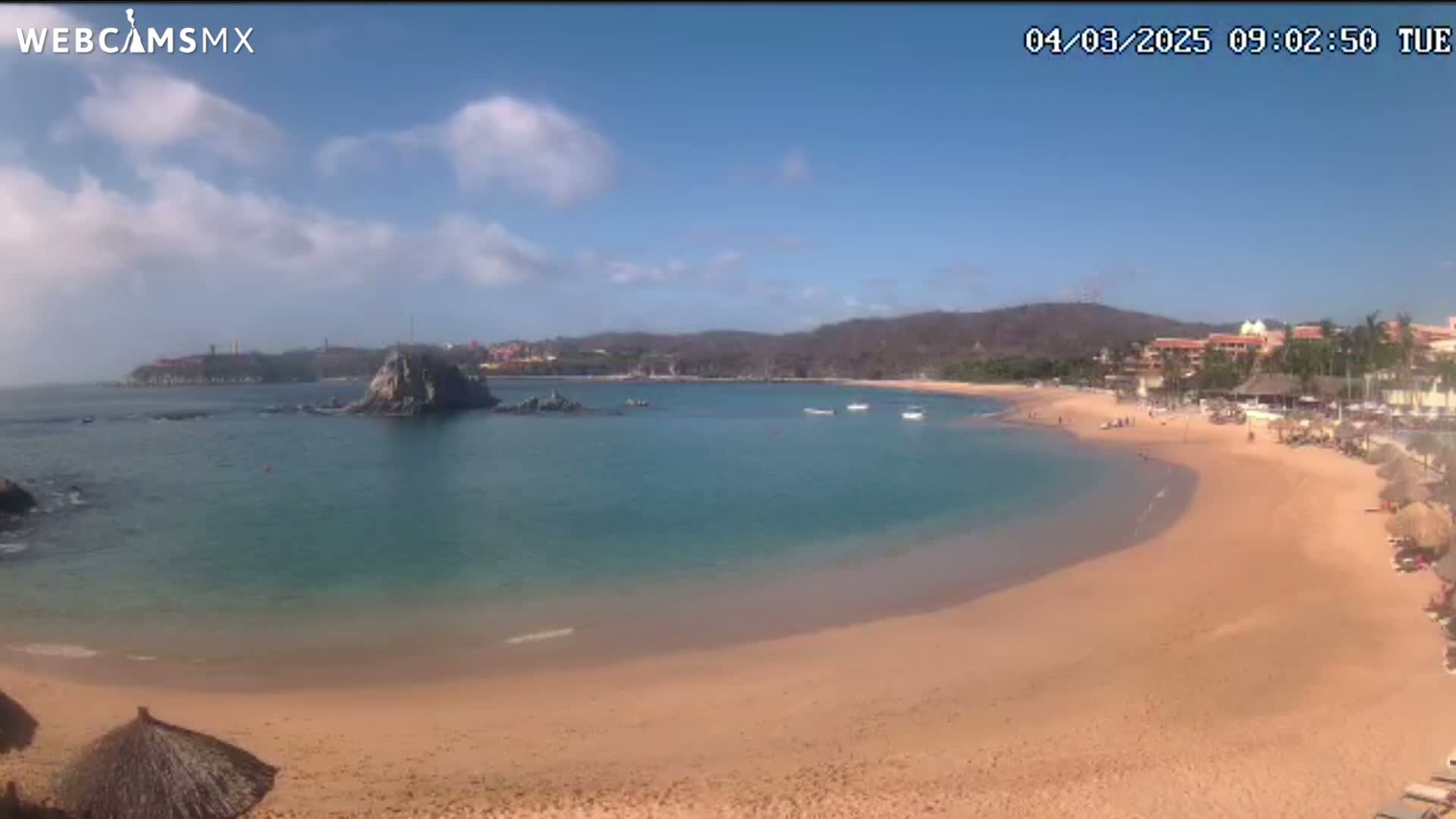 A partly sunny day at a secluded cove with a sandy beach, calm turquoise water, and a rocky outcropping near the shore.
