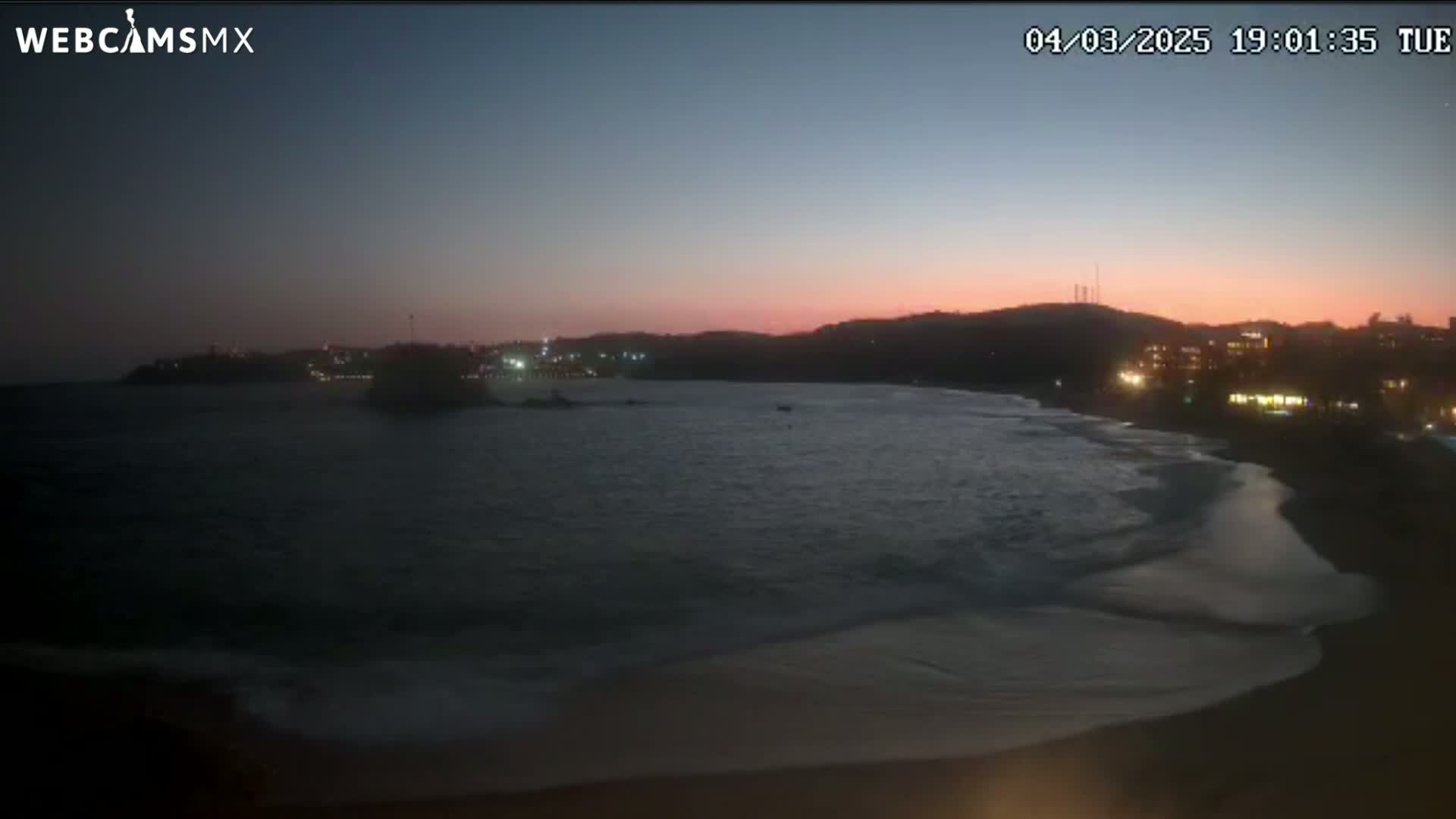 A dark beach at sunset with gentle waves lapping the shore, overlooking a dimly lit town across a calm bay.