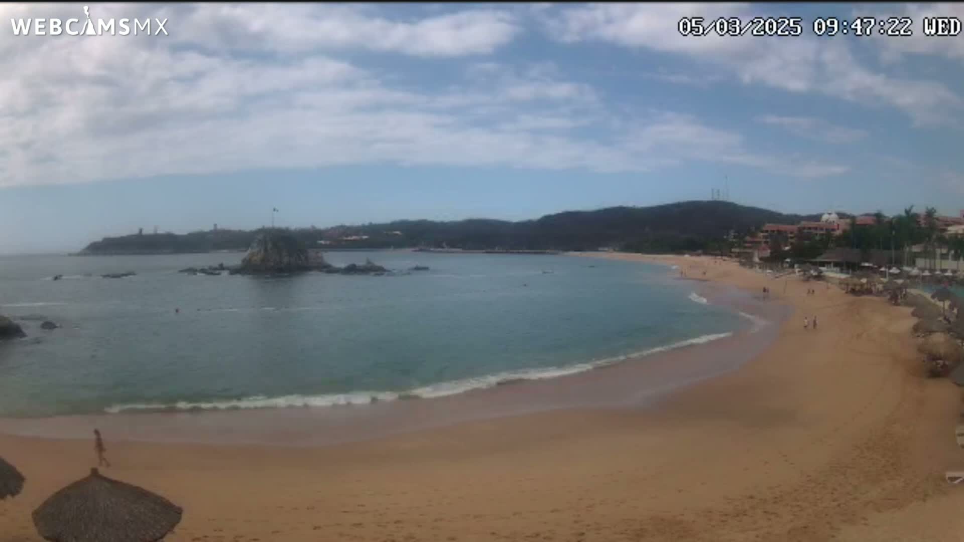 A partly cloudy day shows a sandy beach curving into a calm bay with a rocky island and distant hills.