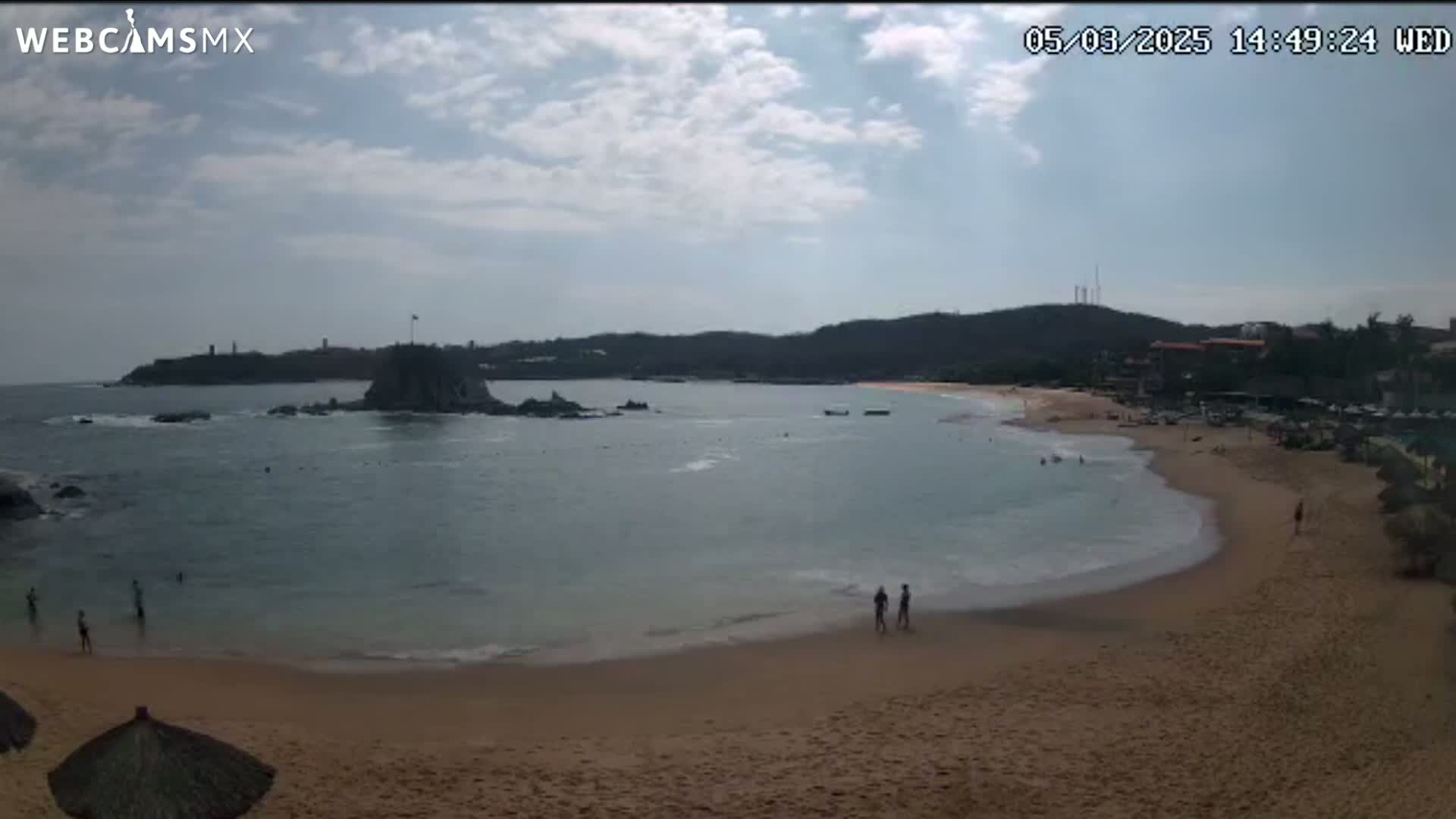 A sandy beach with calm, light-blue water curves around a rocky point under a partly cloudy sky.