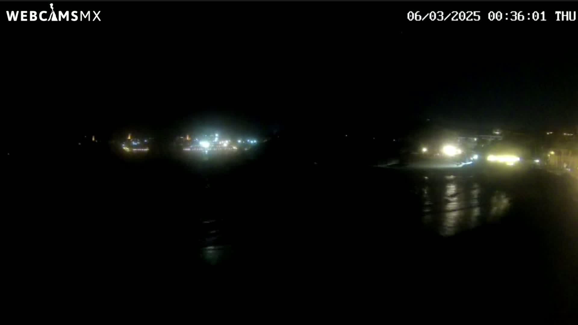 A dark nighttime view of a coastline shows dimly lit buildings and a dark body of water reflecting some light.