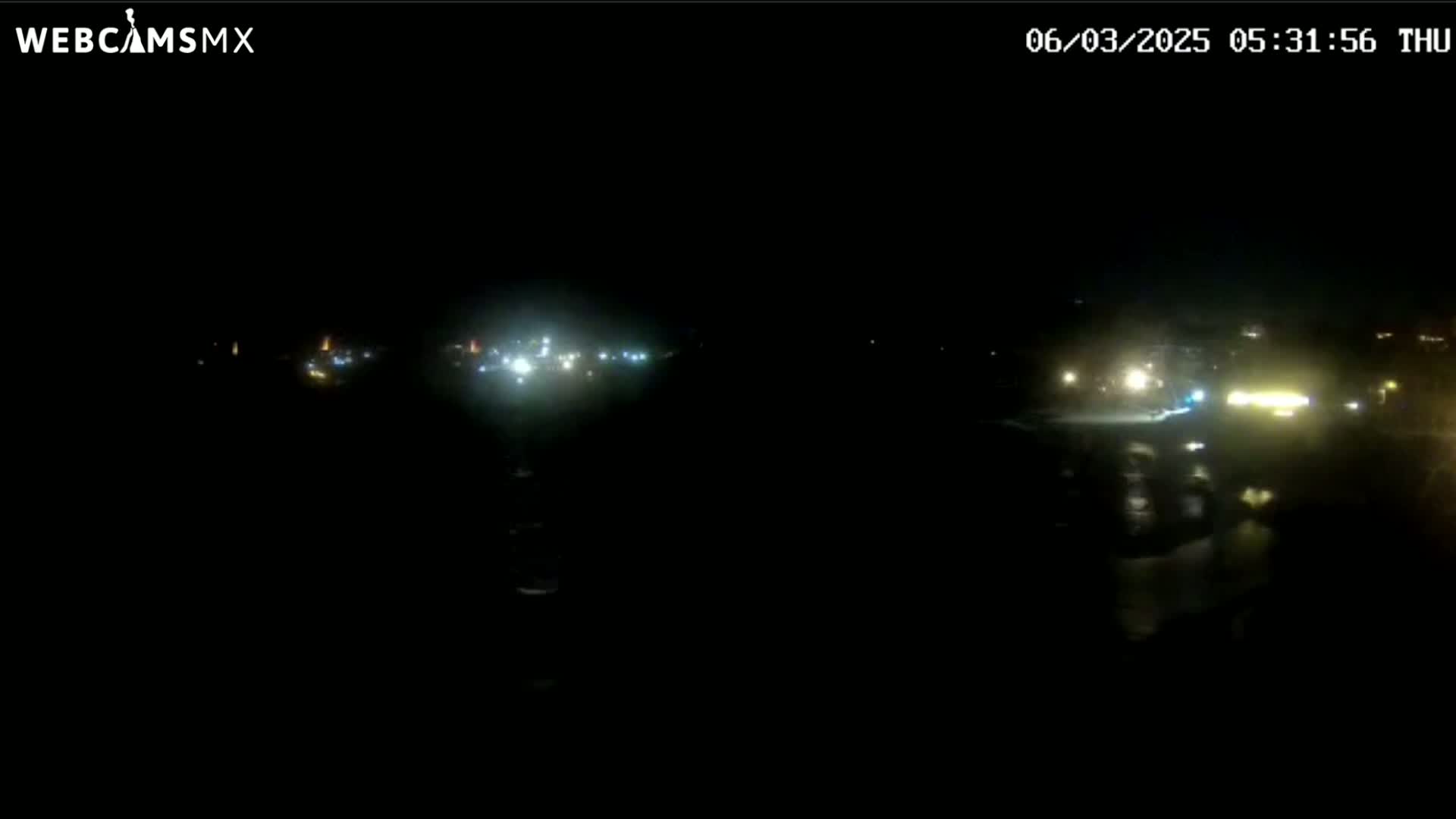 A nighttime long shot shows a dark body of water reflecting faint lights from a distant shoreline.