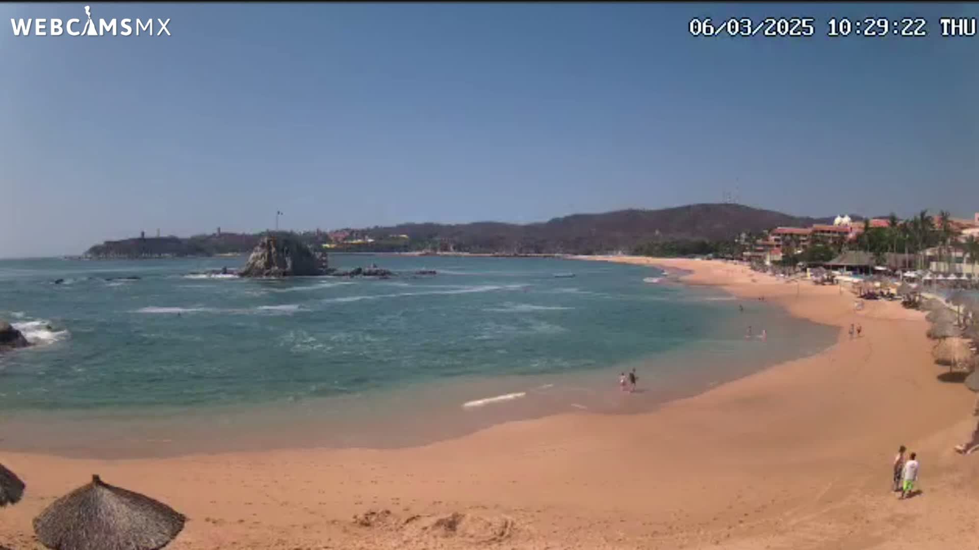 A sandy beach curves along a calm, teal ocean under a clear blue sky, with a rocky outcropping and distant buildings visible.