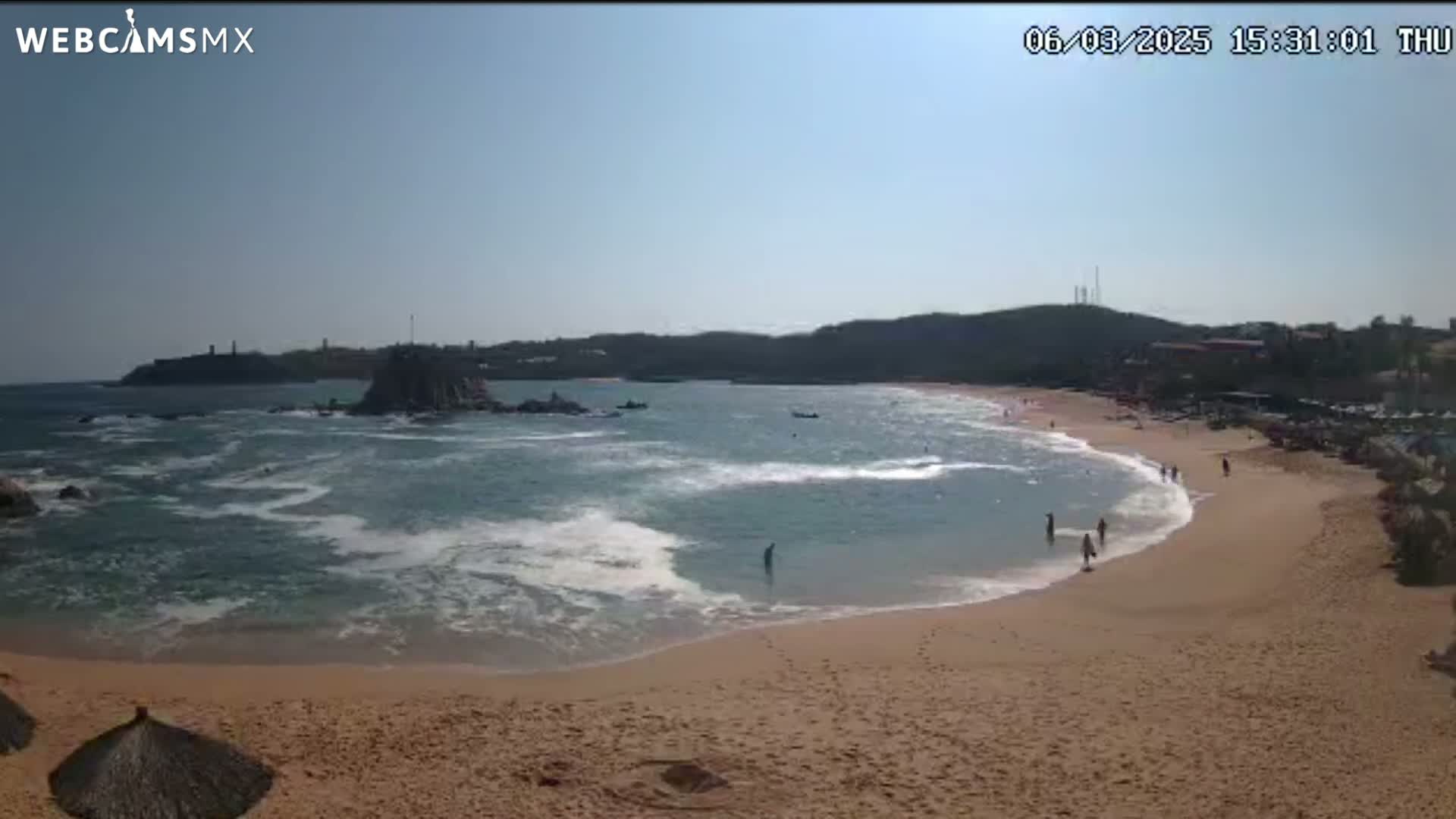 A sandy beach with calm, light blue water, some people swimming and walking, and rocky outcroppings in the bay on a sunny day.