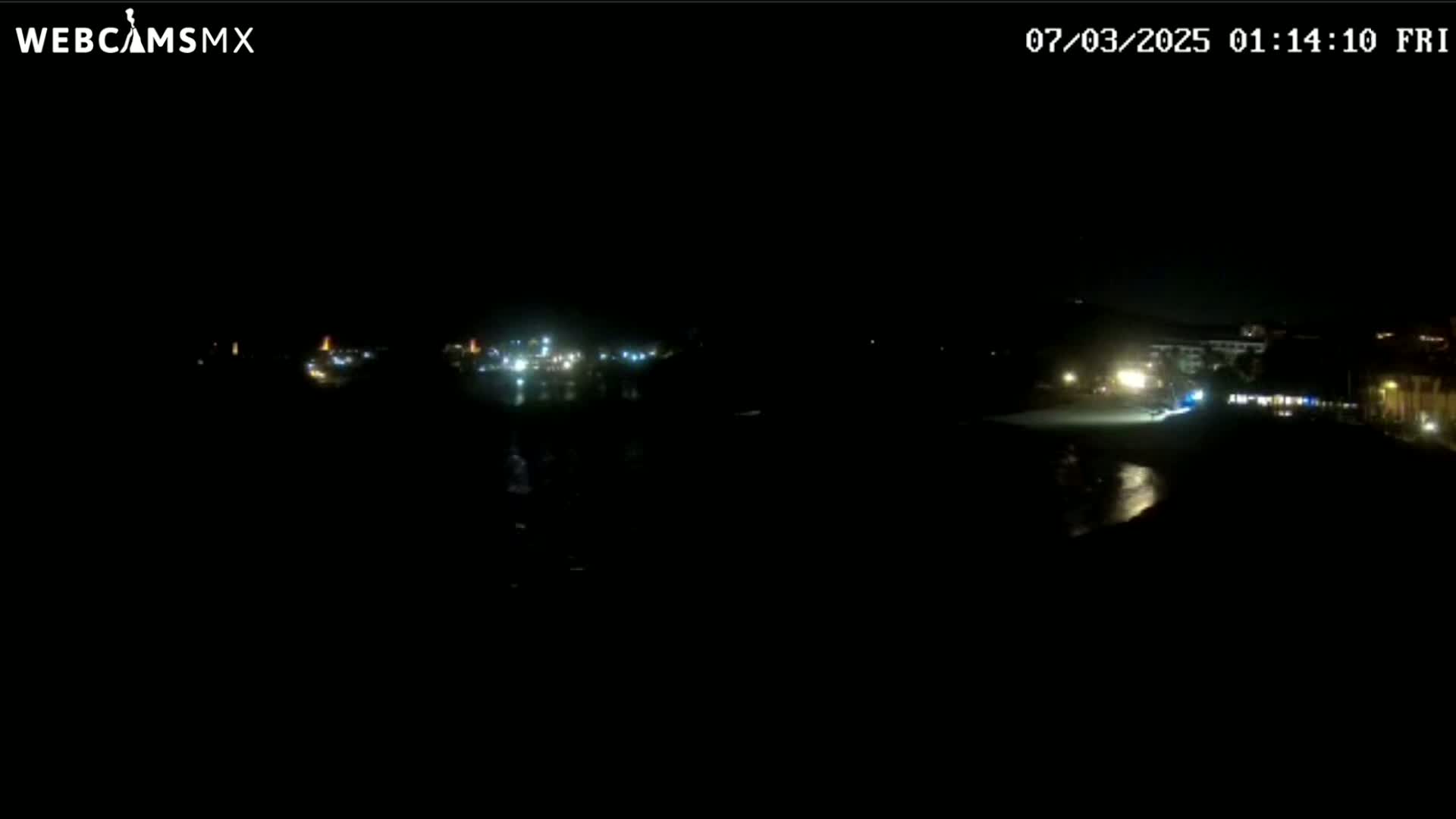A dark nighttime view shows a dimly lit shoreline with buildings and a body of water.