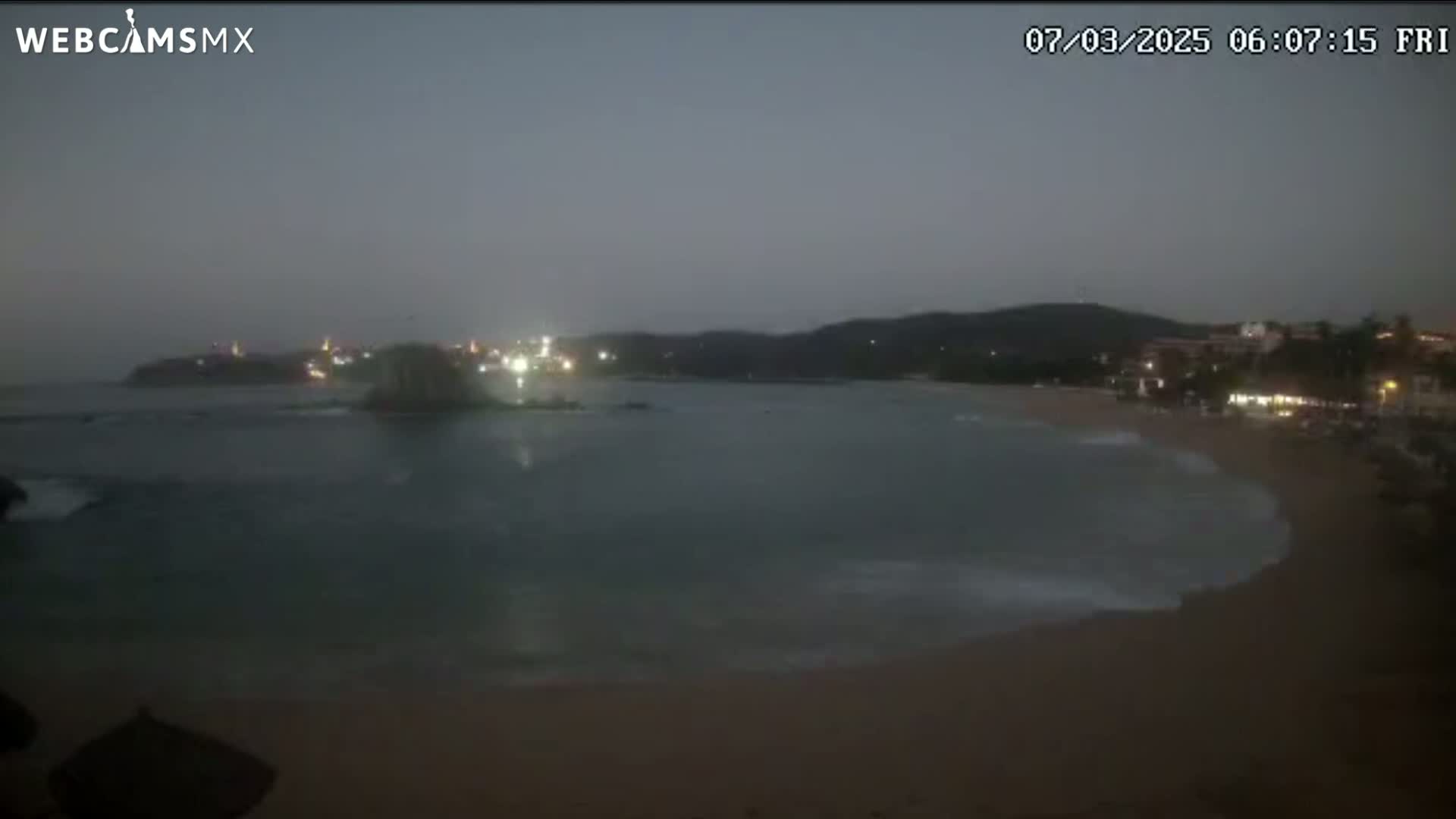 A dark, overcast night scene shows a beach with gentle waves lapping the shore, and a calm bay with dimly lit buildings and vegetation in the distance.