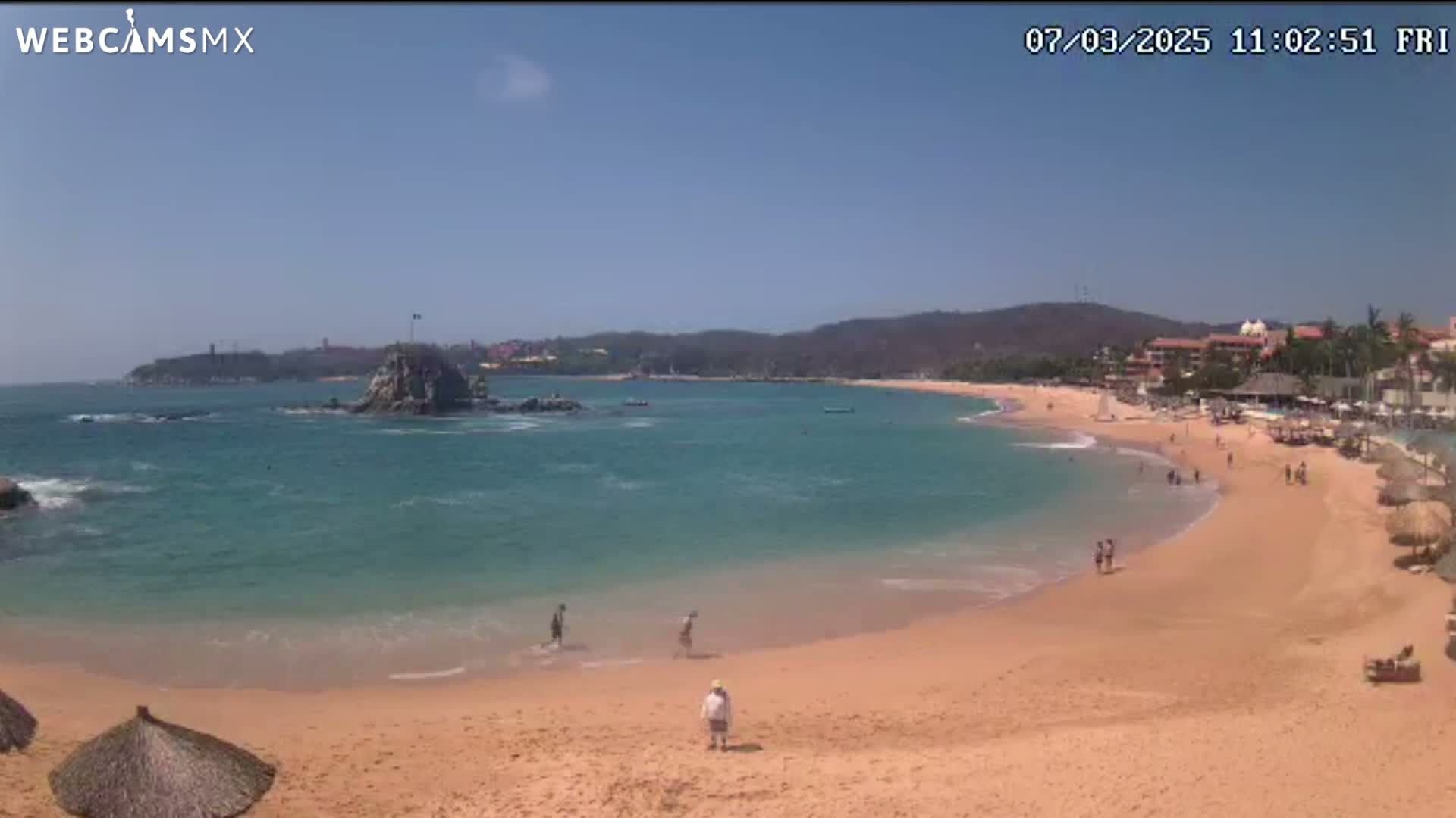 A sandy beach curves around a calm, teal ocean under a clear, sunny sky, with a few people scattered on the sand and in the water, and a rocky outcrop visible in the bay.