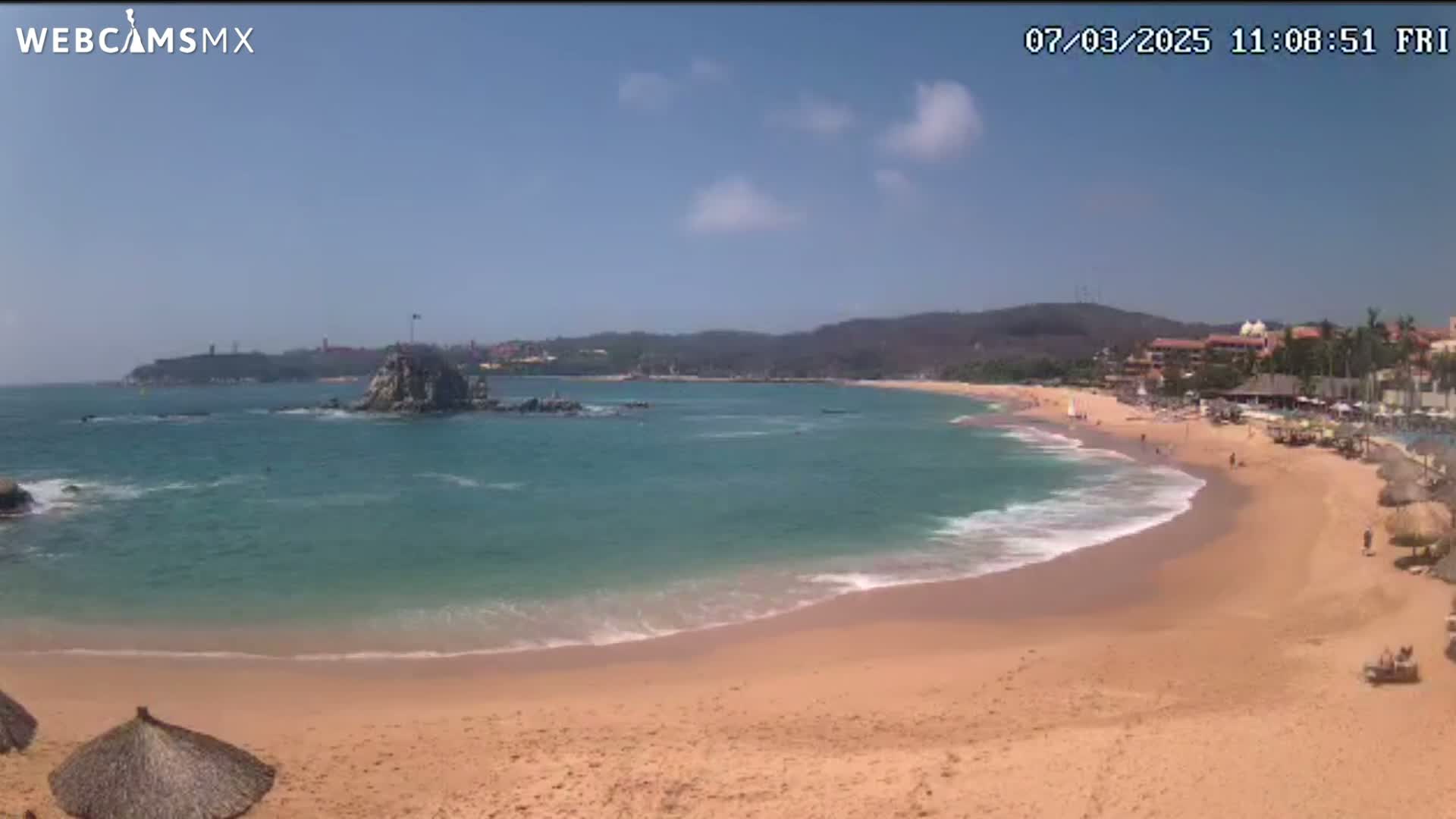 A sandy beach with calm, turquoise water laps gently at the shore under a mostly clear, sunny sky; a rocky outcropping sits in the bay, and buildings are visible in the distance.