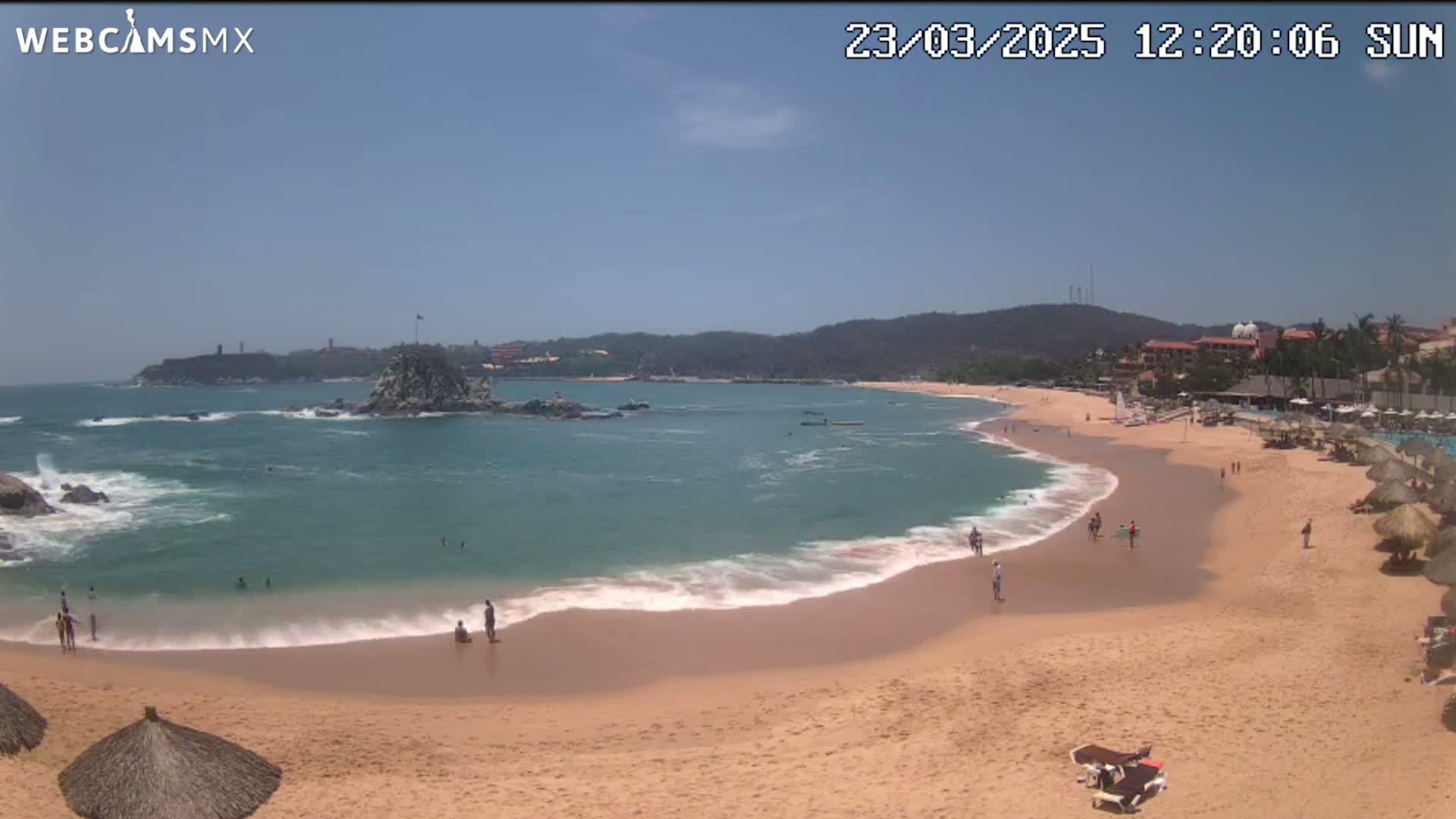 A sandy beach with calm waves laps the shore, under a clear blue sky, with a rocky outcrop and distant buildings visible.