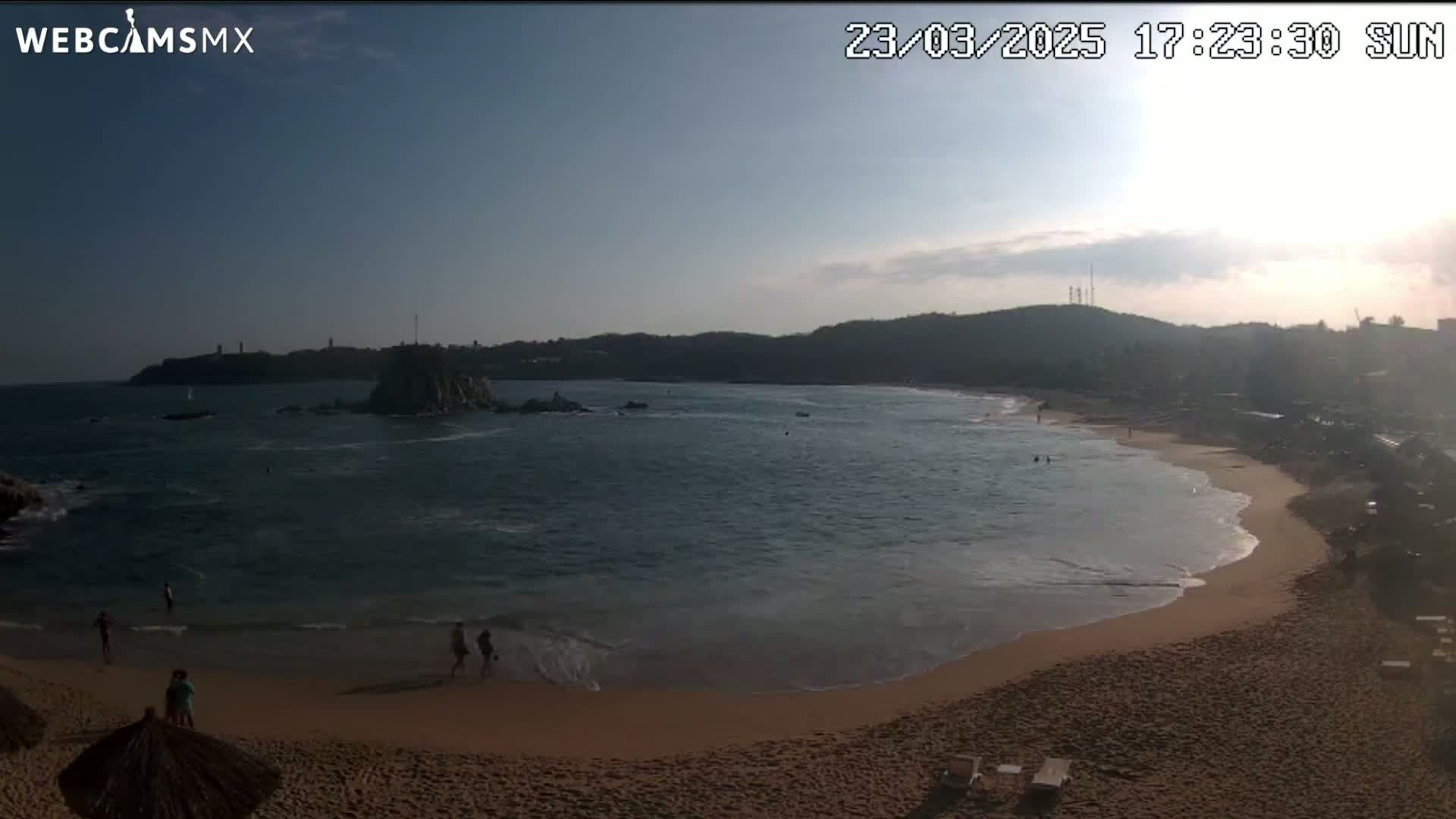 A sandy beach curves around a calm bay under a mostly clear, sunny sky, with a few people visible on the beach and in the water.