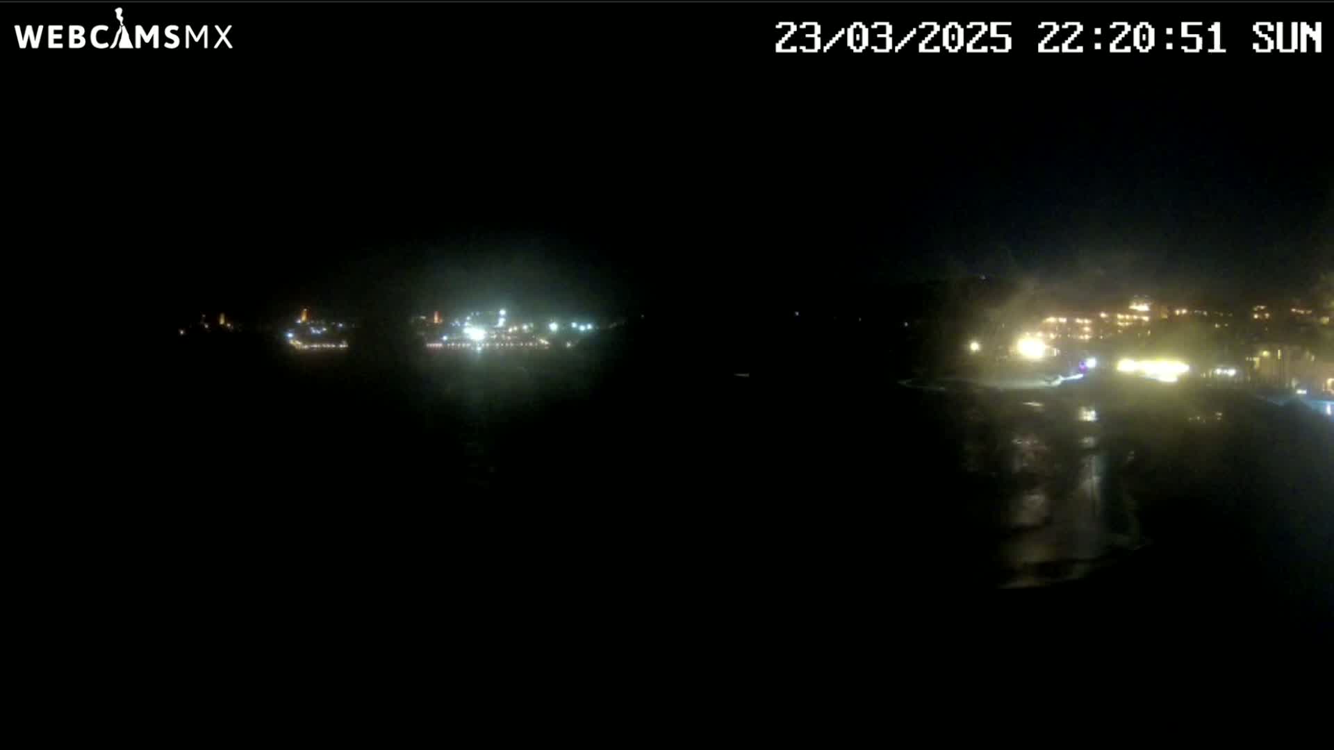 A nighttime long shot of faintly lit buildings and structures across a dark body of water.