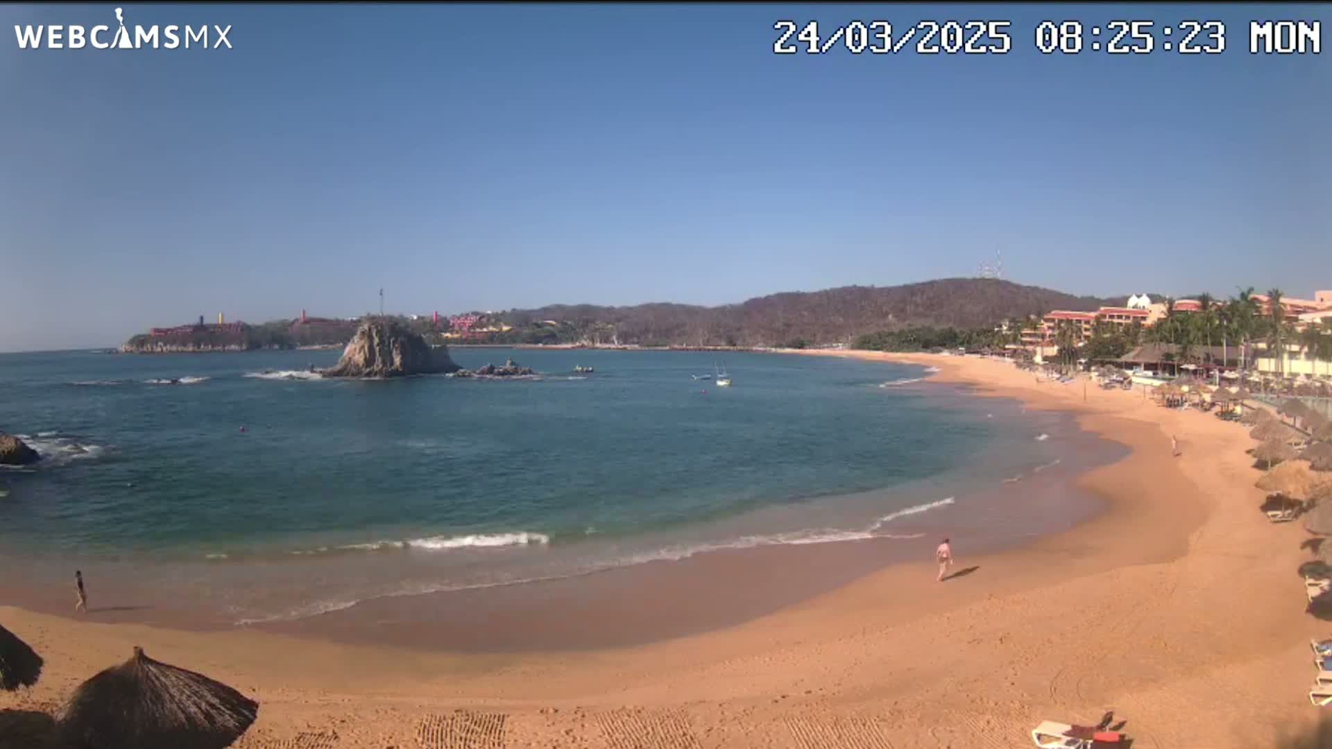 A sandy beach curves around a calm bay under a clear blue sky, with a rocky island and distant buildings visible.
