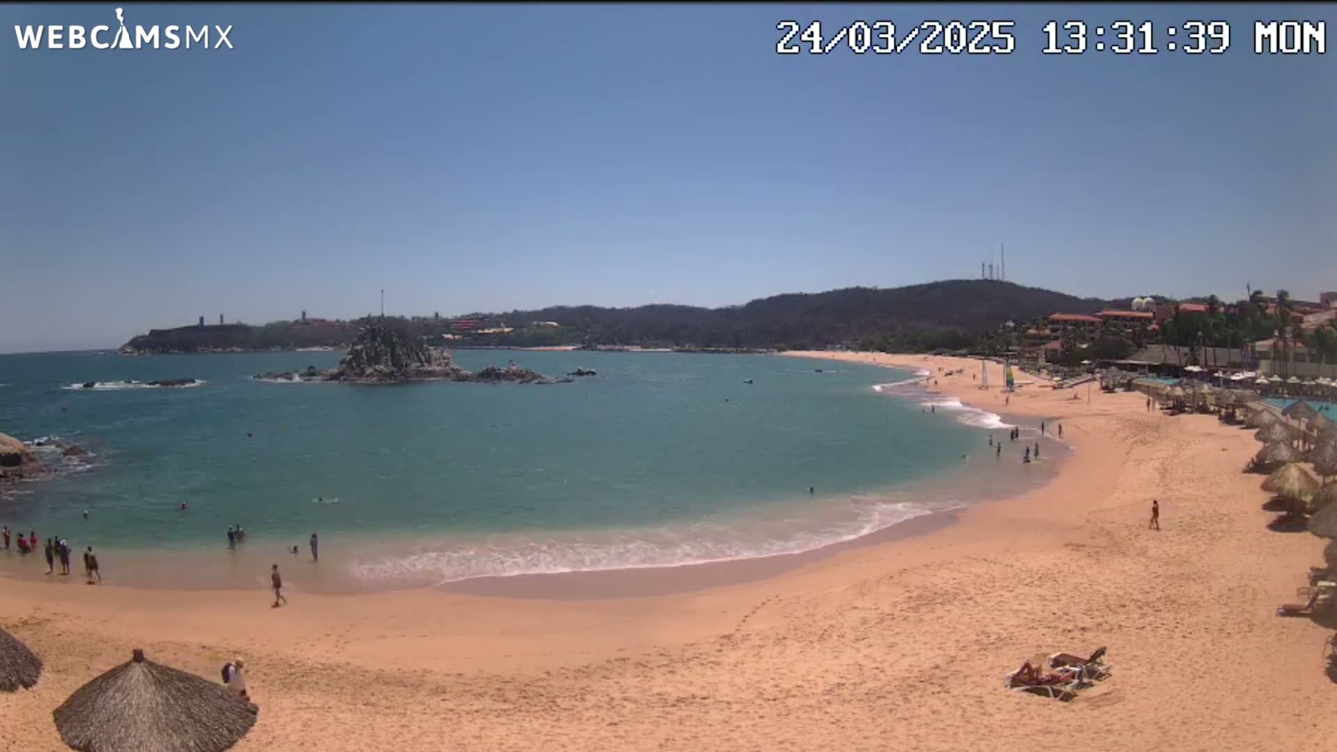 A sandy beach curves around a calm, turquoise bay under a clear blue sky, with people swimming and relaxing on the shore.
