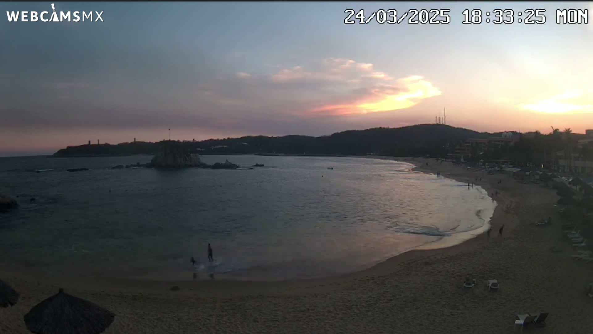 A tranquil beach scene at sunset shows calm water lapping a sandy shore with a few people near the water's edge, and hills in the distance under a partly cloudy sky.