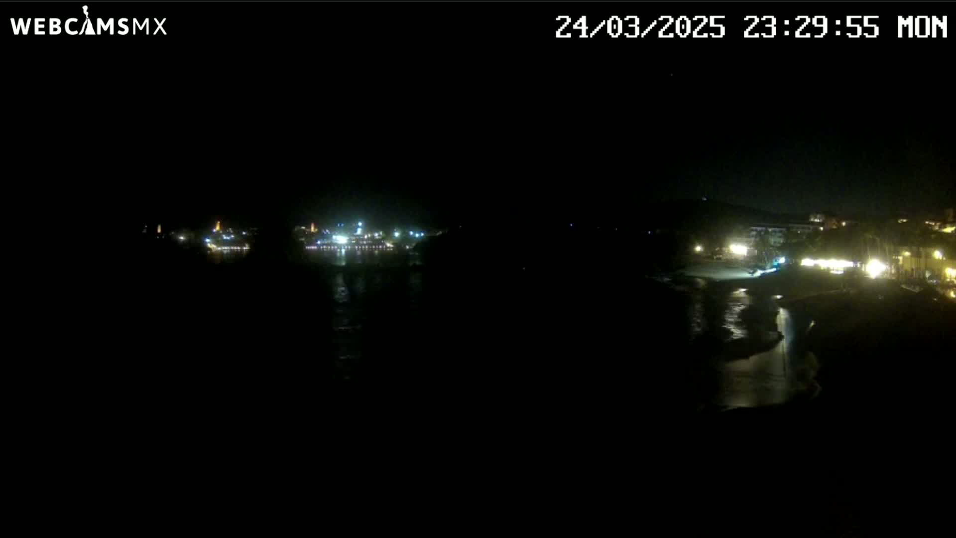 A nighttime view of a dark coastline with dimly lit buildings and a body of water reflecting some lights.
